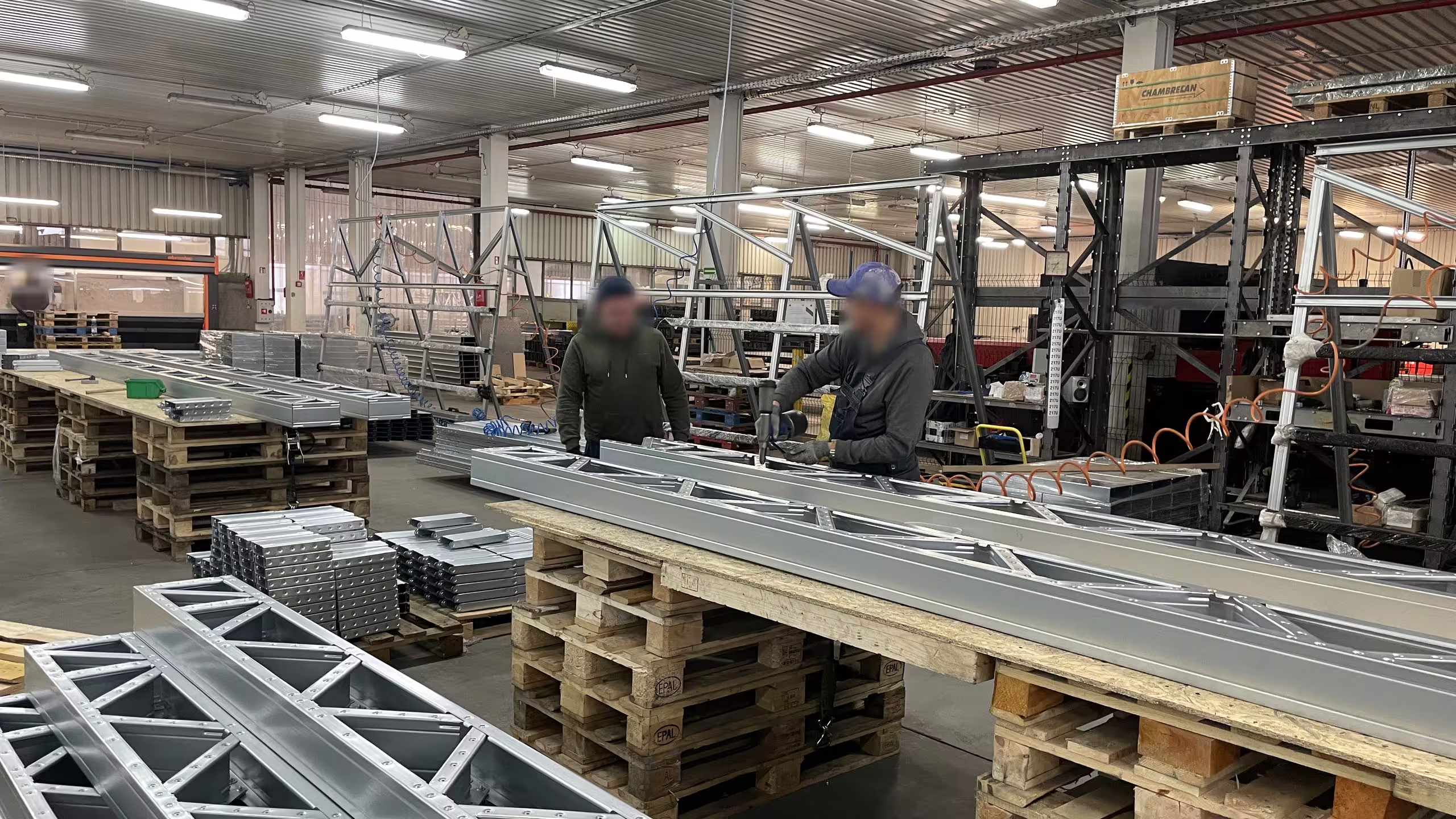 Two fitters bolting up a long lattice beam supported on timber trestles in the Słupy shop, with completed lattice sections stacked in the foreground and racking behind