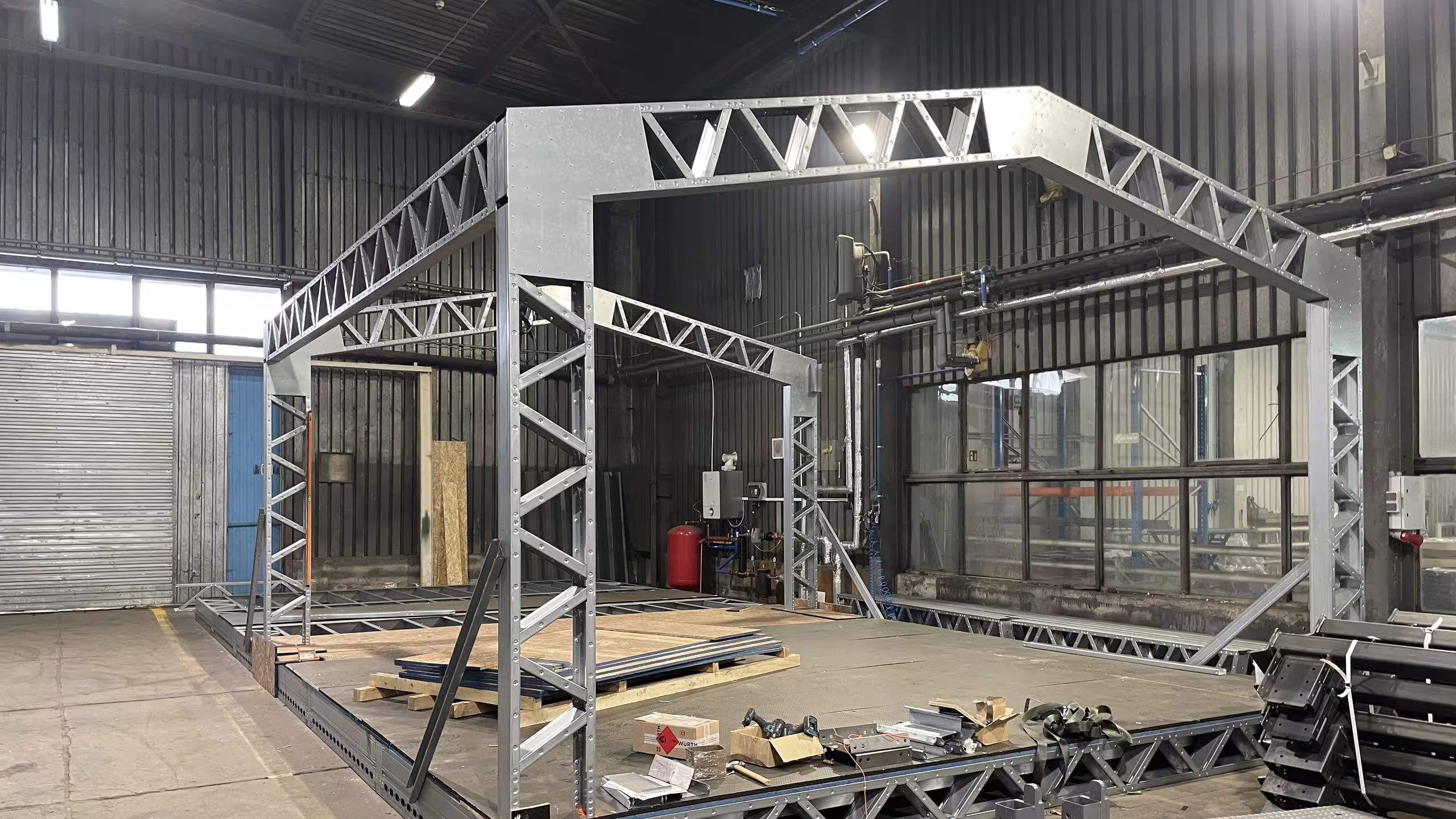 A complete structural bay proof-assembled inside the Słupy works: lattice portal frames and ridge trusses bolted up over a deck, with loose sections and fixings laid out on the floor alongside