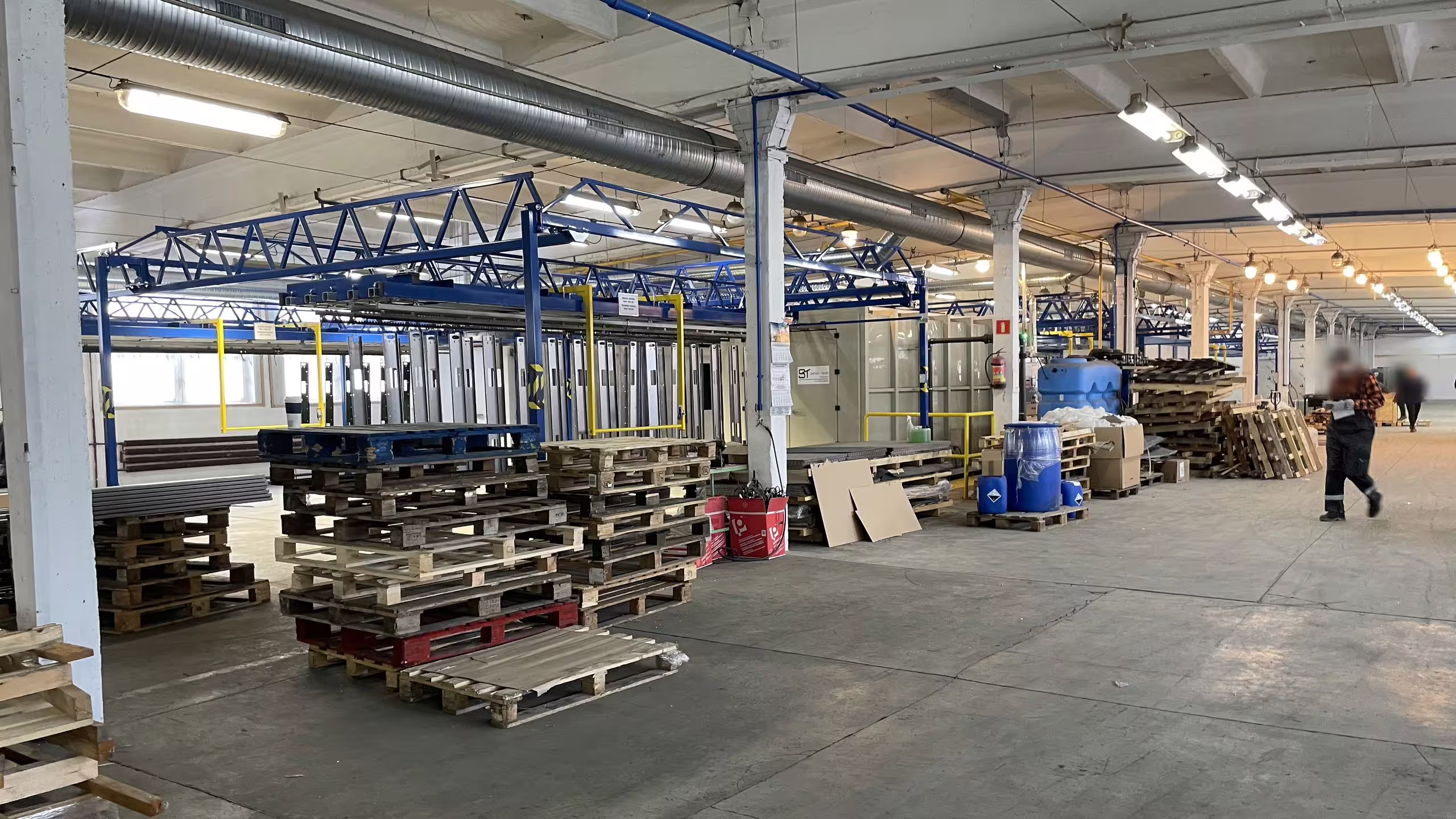 The main production hall at Słupy under its blue overhead crane gantry, with stacked pallets and staged panel assemblies down the left side and open floor running the length of the bay