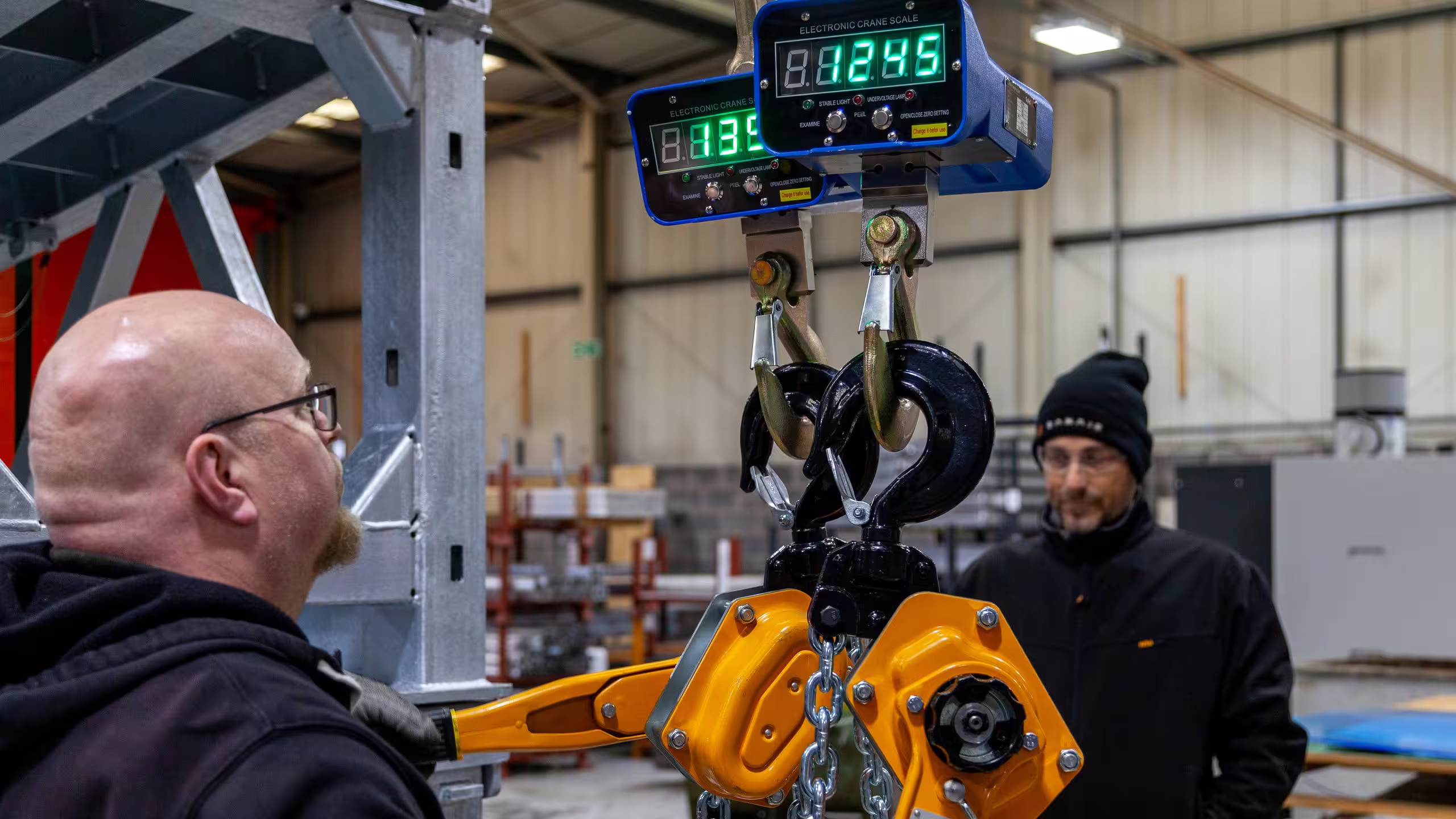 Two BANAIR engineers in a UK workshop during a load test: one in the foreground watching a digital crane scale hanging from chain hoists, the second standing beside the rig in a dark work jacket