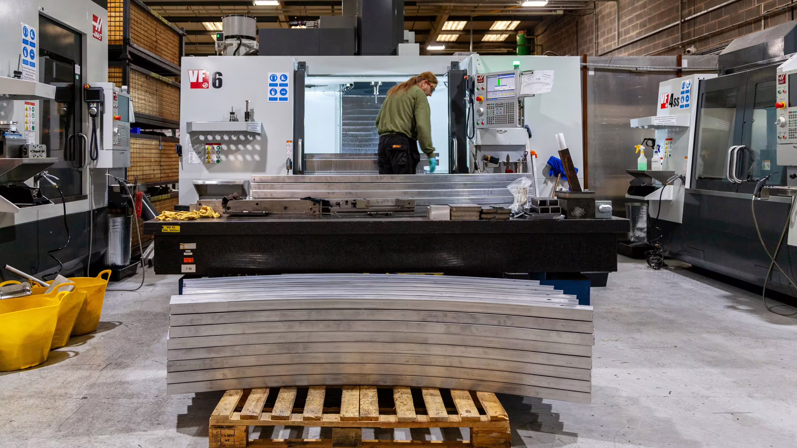 A machinist working inside a CNC machining centre in the BANAIR UK plant, flanked by two further machining centres, with a long aluminium section set up on the machine bed and a stack of curved aluminium profiles on a timber pallet in the foreground