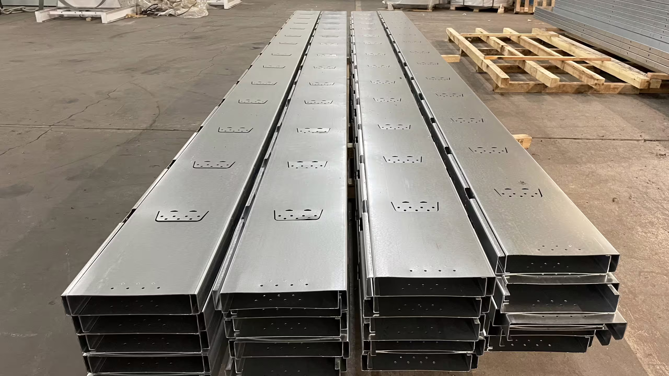 Four runs of roll-formed galvanised C-section stacked on the shop floor at Słupy, each length punched and pre-drilled to the same pattern, with coil stock and a roll-forming line behind