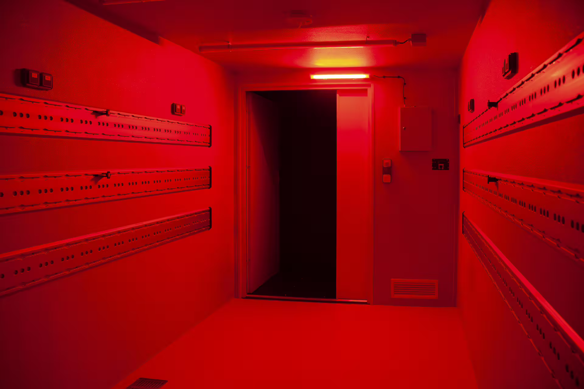 FlexiPod interior under red blackout lighting, showing rail-equipped walls and an open doorway.