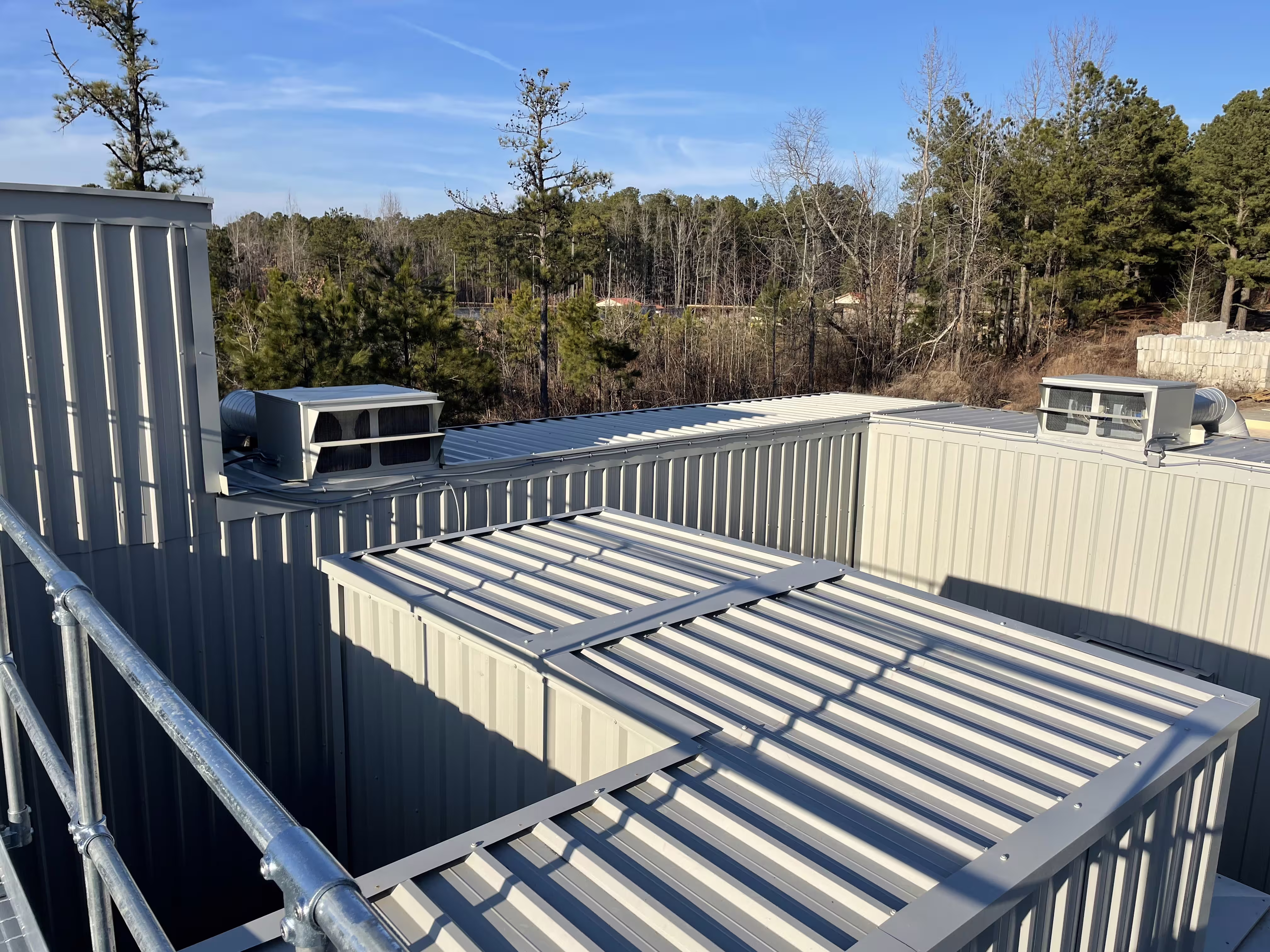 Roof-level view across the MOTC complex: stepped ribbed-steel modules, roof-mounted air-conditioning units and a galvanised walkway guardrail, pine woods behind
