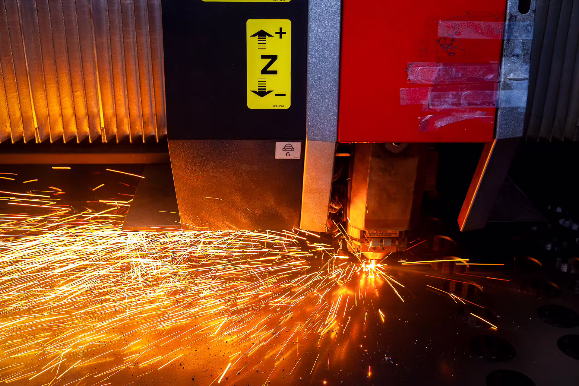 Close-up of a CNC laser cutting head cutting through steel sheet, sending a shower of orange sparks across the plate, with circular holes already cut beside it