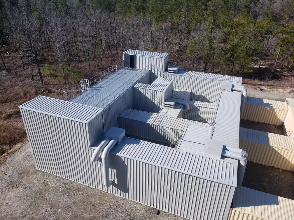 Aerial view of a MOTC urban training complex: interlocking ribbed-steel modules forming a warren of rooms and roof levels, an external stair at the near corner, set on bare ground against winter woodland