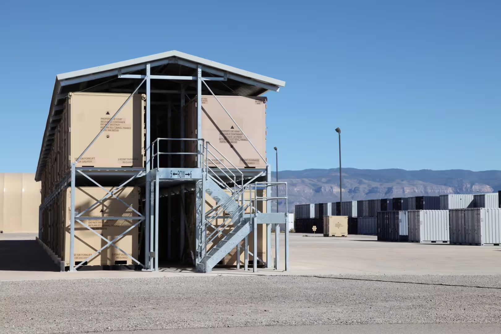 Gable end of a two-storey container racking structure with tan USAF containers and galvanised access stairs at a desert container yard