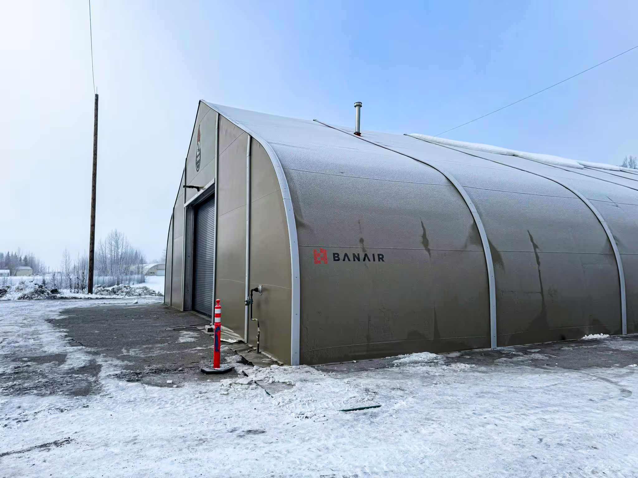 A tan fabric-membrane shelter carrying BANAIR branding on its flank, standing on snow-covered hardstanding under an overcast winter sky, with a roller shutter door open at the gable end, a flue on the roof and an orange delineator post beside the entrance.