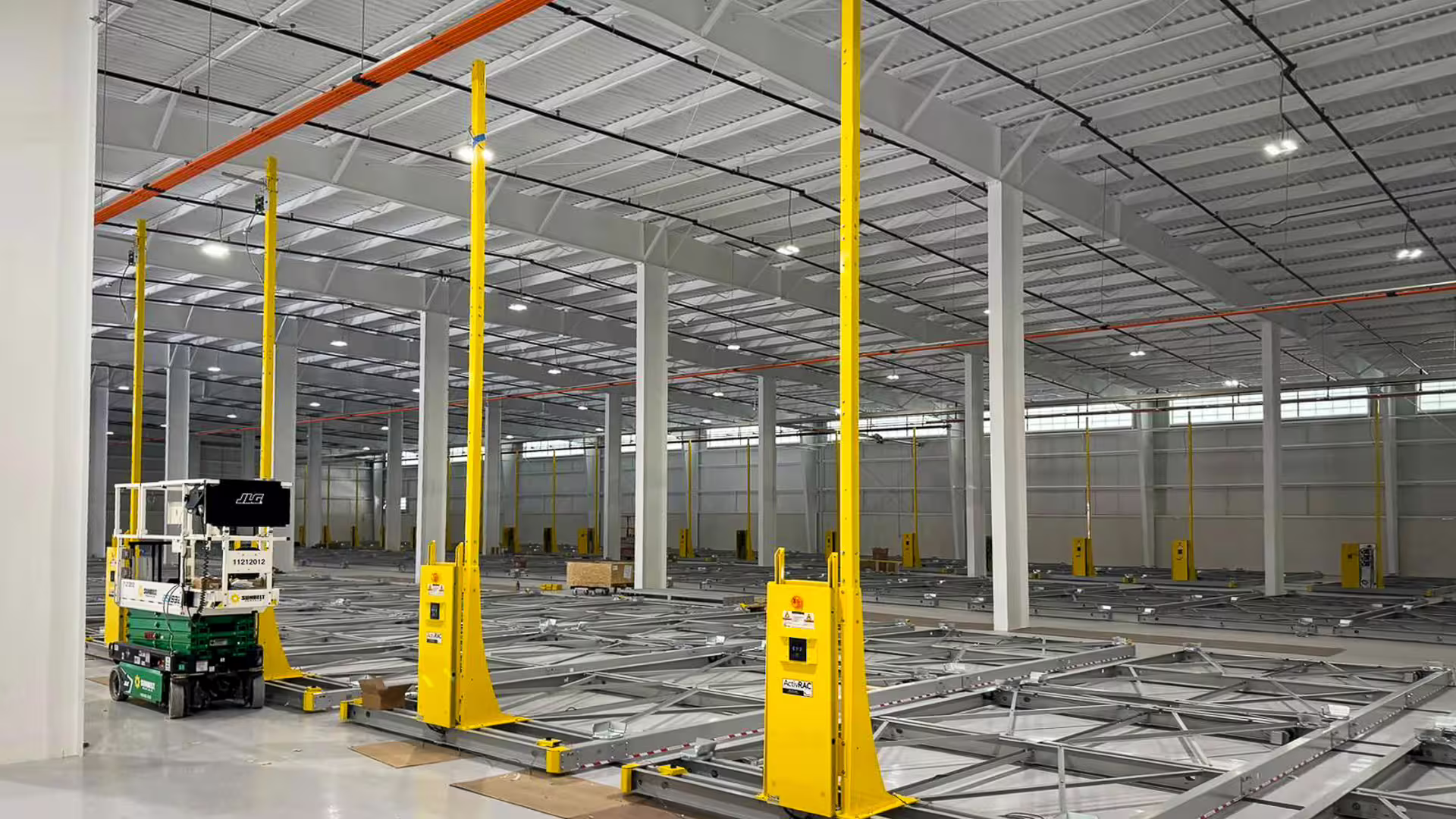 An empty steel-framed hall with RollingStack mobile-base rails set into the floor and yellow guide posts standing at the ends of each run, a small work platform at the left