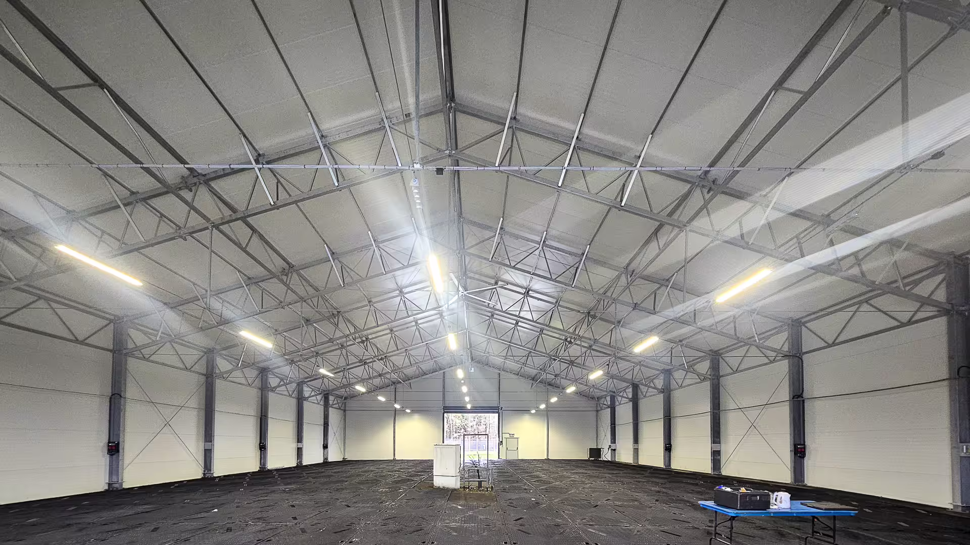 The interior of a DMTSS shelter looking down its length: a clear unobstructed span under a lined roof with continuous lighting runs, roller-shutter doors in the end wall and a bare floor