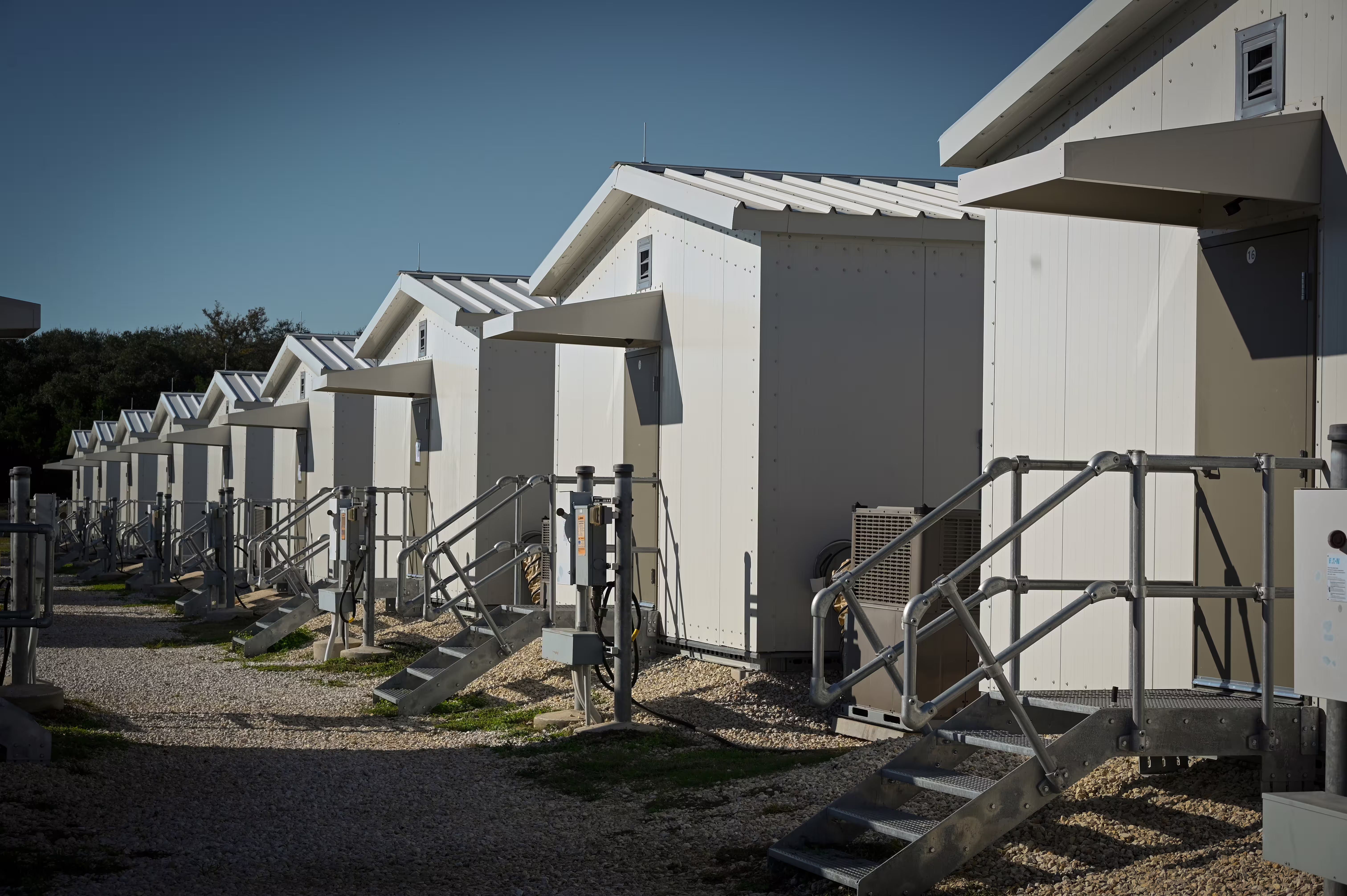 A row of white Tactical Shelters with pitched metal roofs and door canopies, each with galvanised steel entry steps, an external air-conditioning unit and a power pedestal, on a gravel site under a clear sky