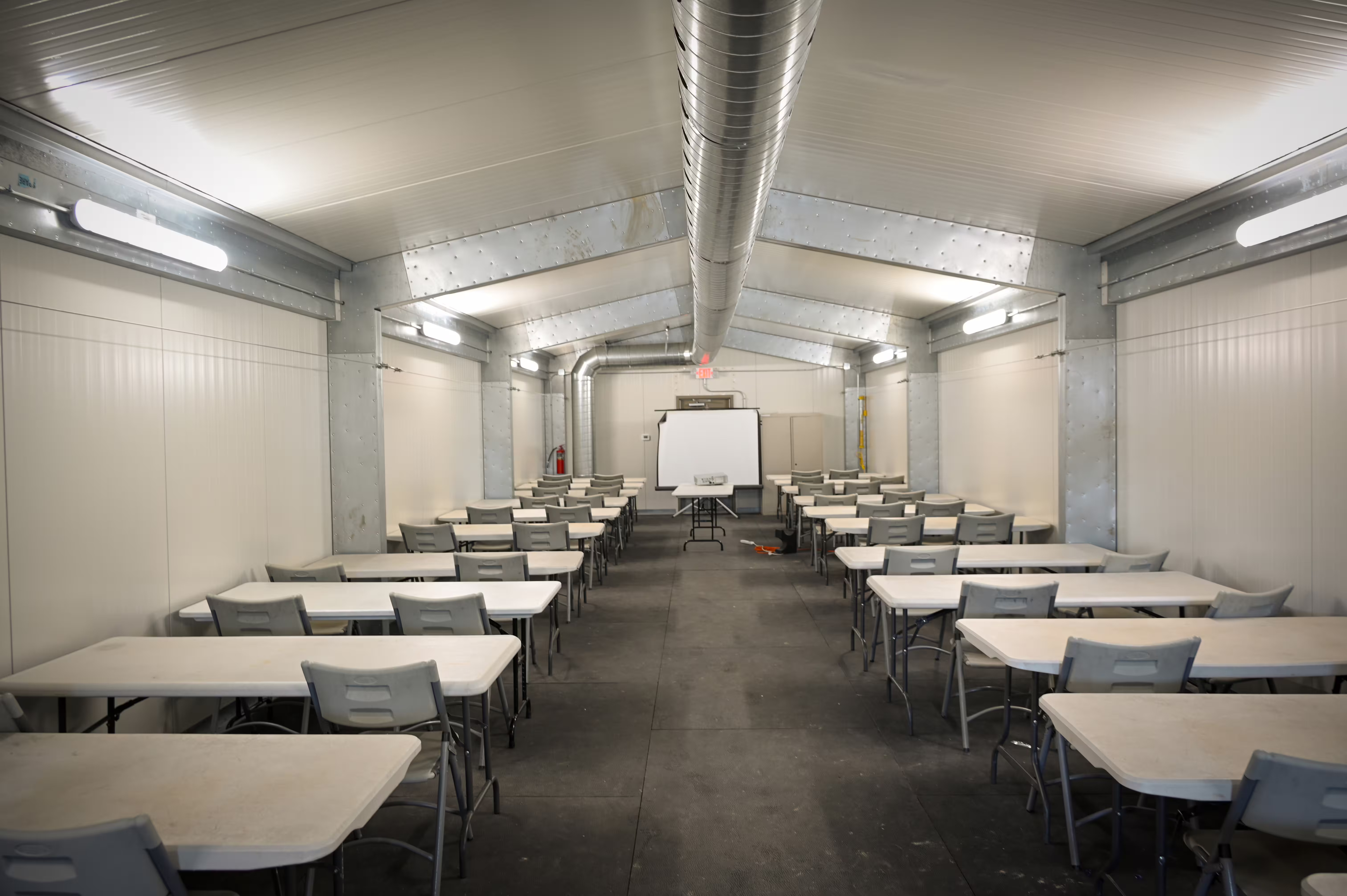 The finished interior of a Tactical Shelter: white insulated wall and ceiling panels, continuous ceiling lighting, a dark floor, a personnel door at the far end and a wall-mounted environmental unit