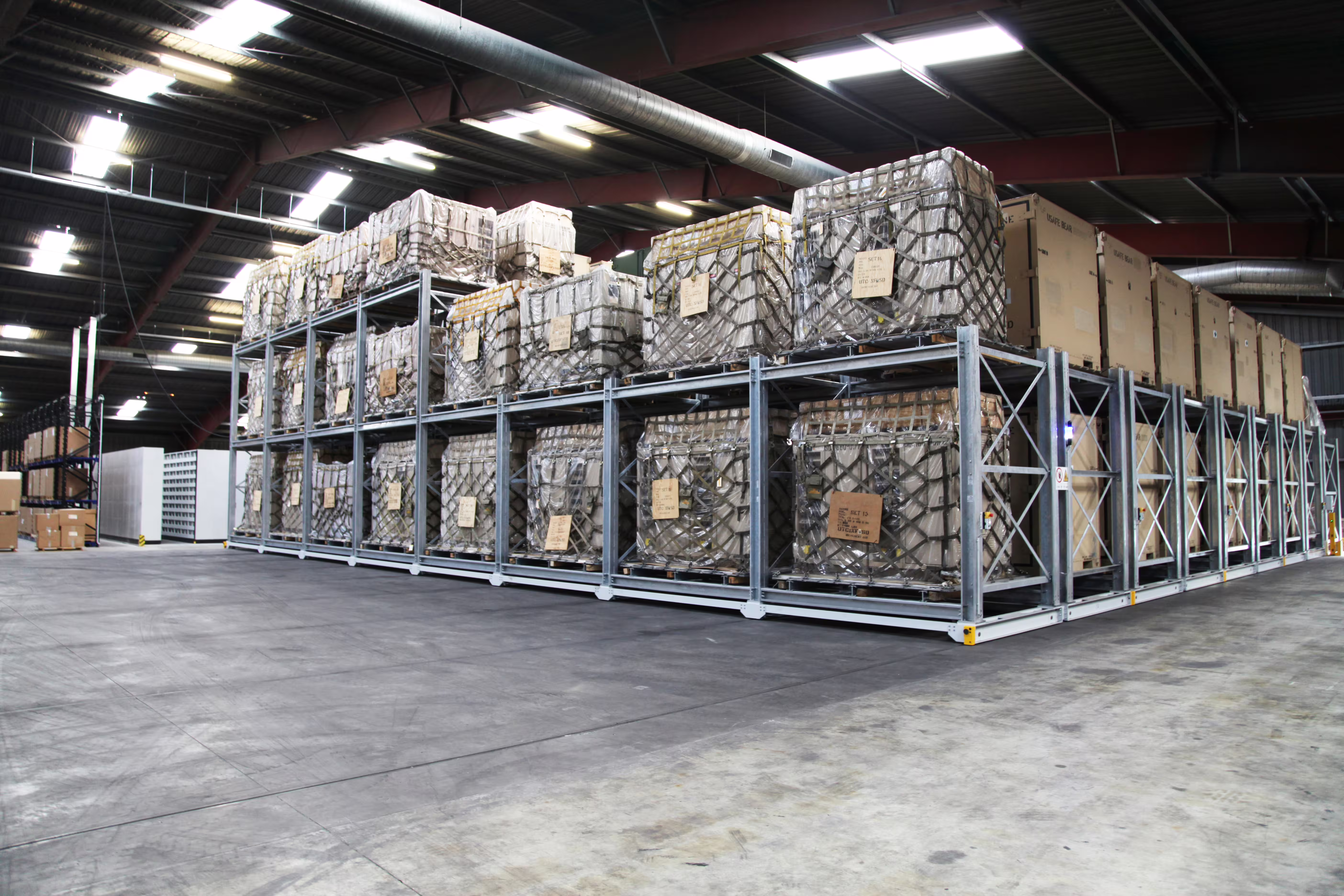 A large block of galvanised CargoRack bays on mobile bases inside a hangar, rolled together without aisles and loaded to three levels with palletised and netted stores