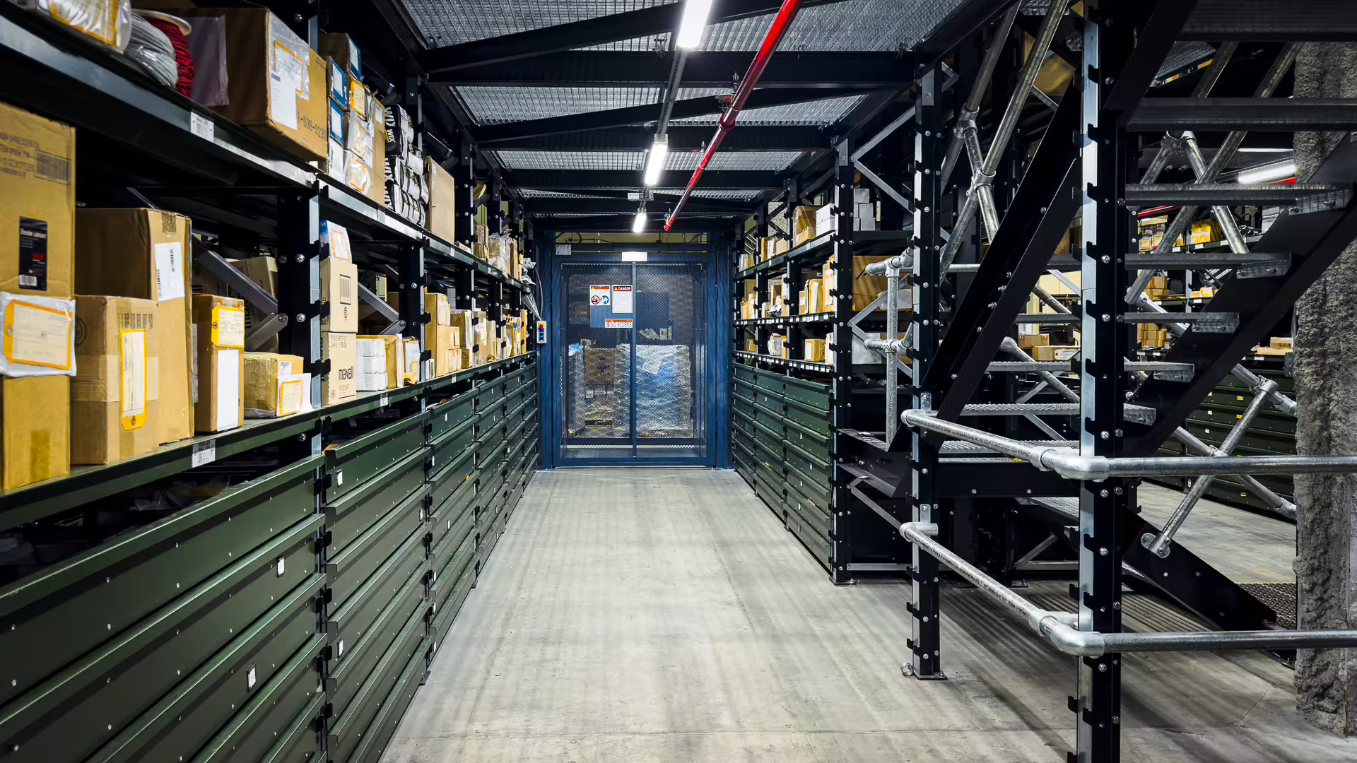 A warehouse aisle lined with green RackMezz drawer units, with a bolted steel staircase and tubular handrail climbing to the mezzanine deck on the right