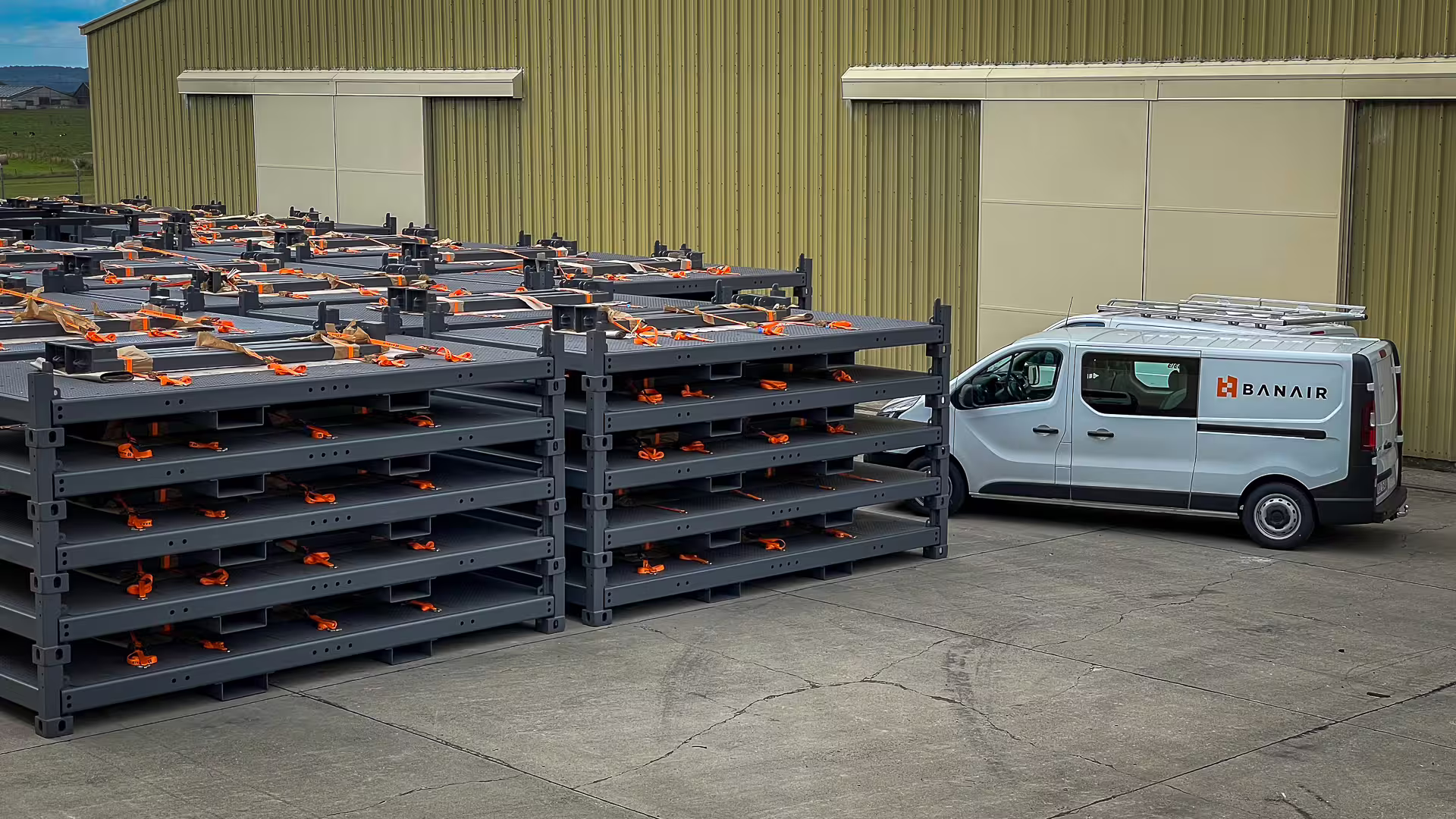Two stacks of empty RollingStack frames standing in a yard, five and six levels high with their corner posts seated into each other and orange lashings across the decks, a BANAIR van alongside for scale