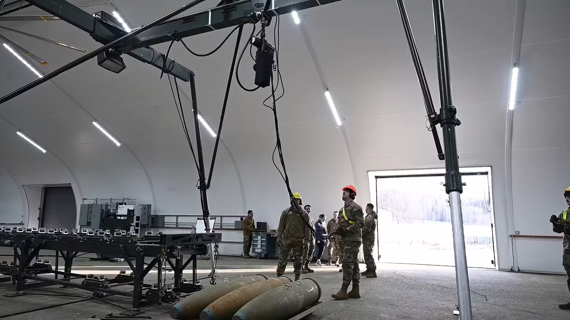 Personnel in hard hats rigging an overhead gantry hoist inside a fabric-membrane shelter, with a roller conveyor line along one wall, large ordnance bodies on the floor and an open roller door showing snow outside.