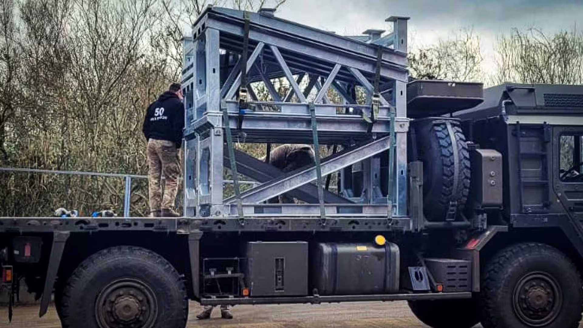 A galvanised MUDR-PD90 ramp section loaded on the flatbed of a military truck, its lattice frame strapped down, with a member of staff standing alongside
