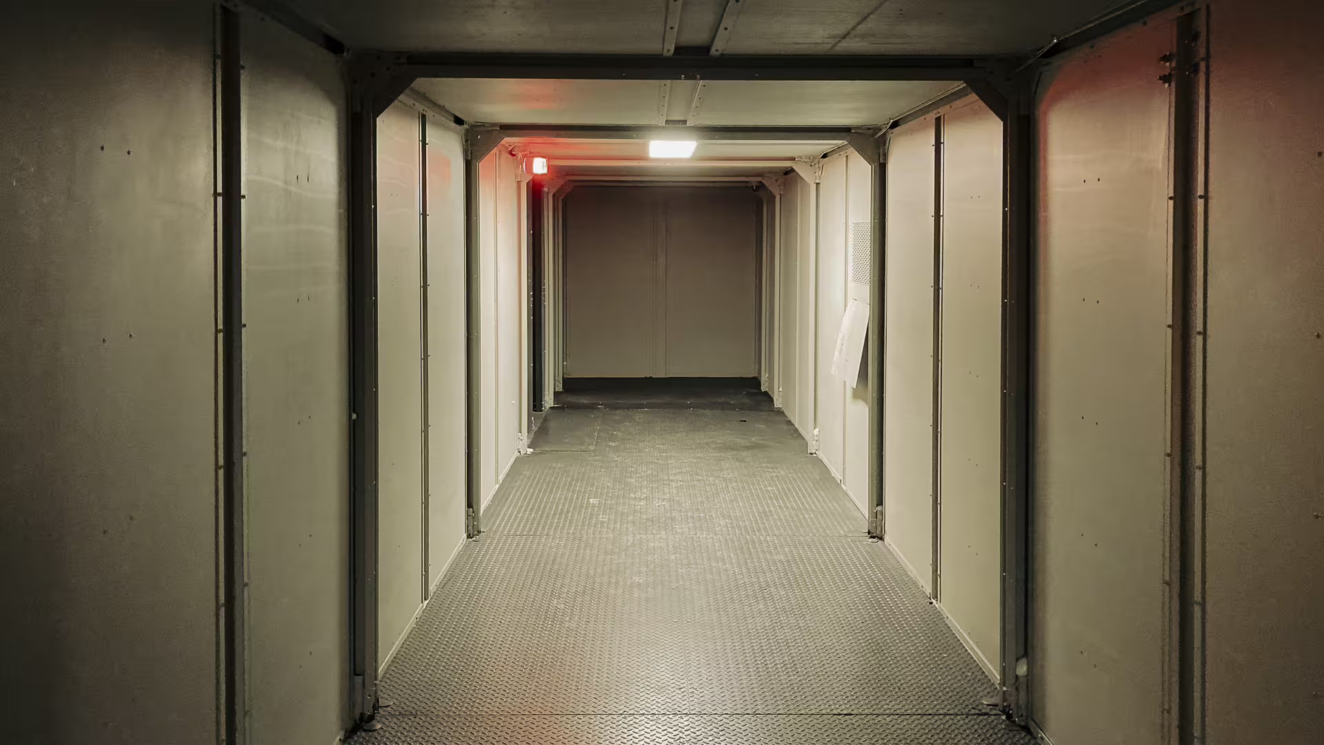 A corridor running the length of a MOTC module: panel walls and door openings on both sides, chequer-plate floor, ceiling lighting overhead and a red lamp burning at the far end