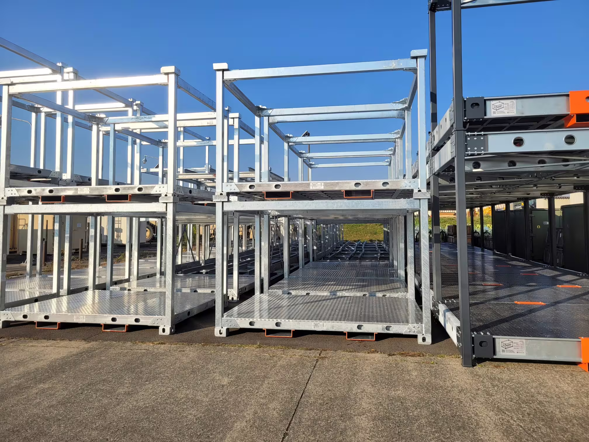 Two-level galvanised open-frame racks standing on an apron, their mesh decks bare and orange corner markers fitted, with further racks lined up behind