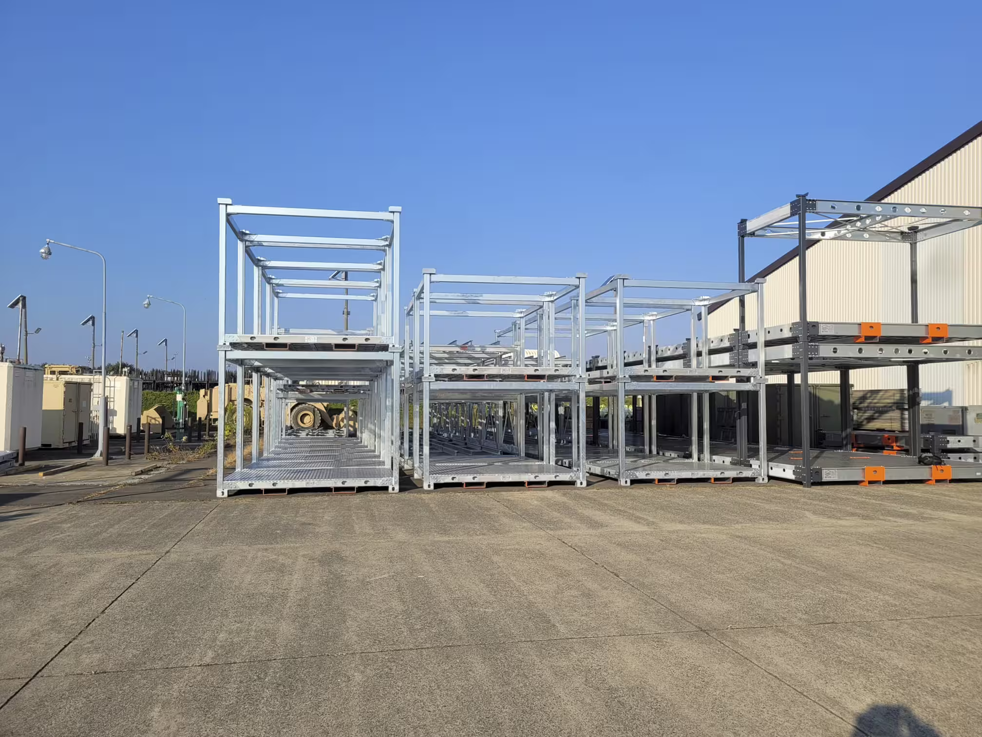 Rows of galvanised two-level crate frames lined up on an apron three bays deep, with low buildings and lighting masts on the skyline behind