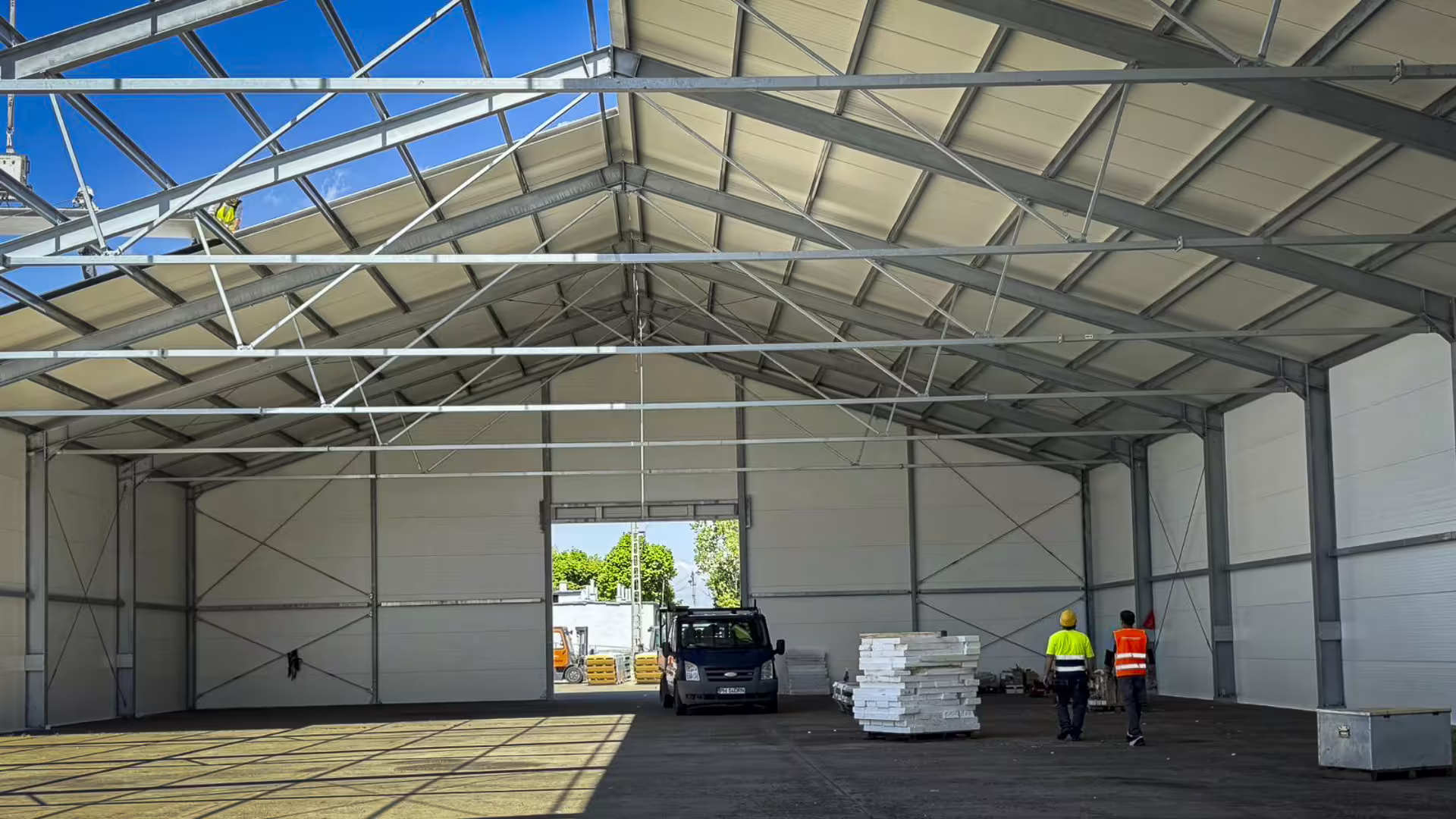 Inside a DMTSS building during assembly: a crew clads the roof between galvanised steel frames while two workers in hi-vis walk past pallets of panels, with a van parked by the open doorway