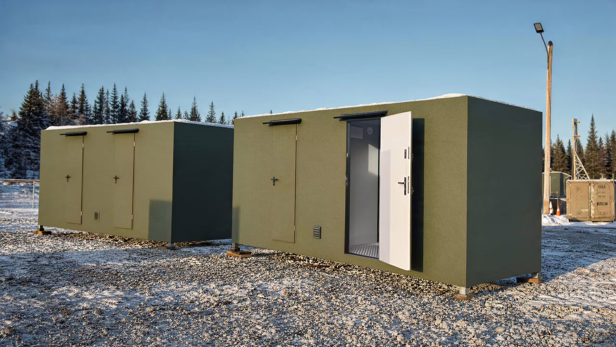 Visualisation of two BunkBox units standing in line on snow-covered gravel in low winter sun, flank doors and ventilation louvres along the near elevation, conifers and a lighting column behind