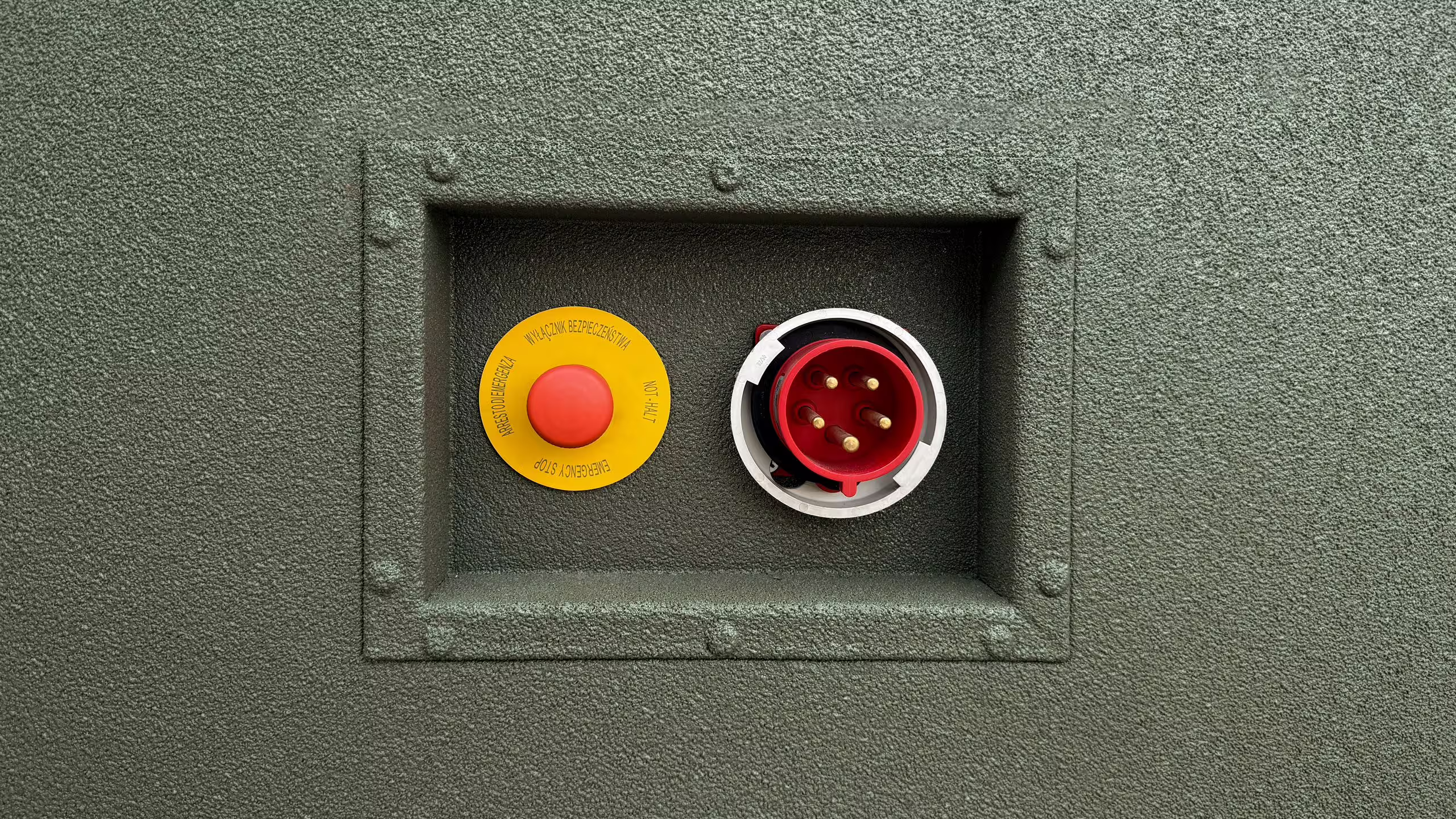 A recessed service panel in the green wall of a BunkBox unit, holding a red five-pin industrial mains connector and a yellow-and-red emergency stop button behind a riveted frame