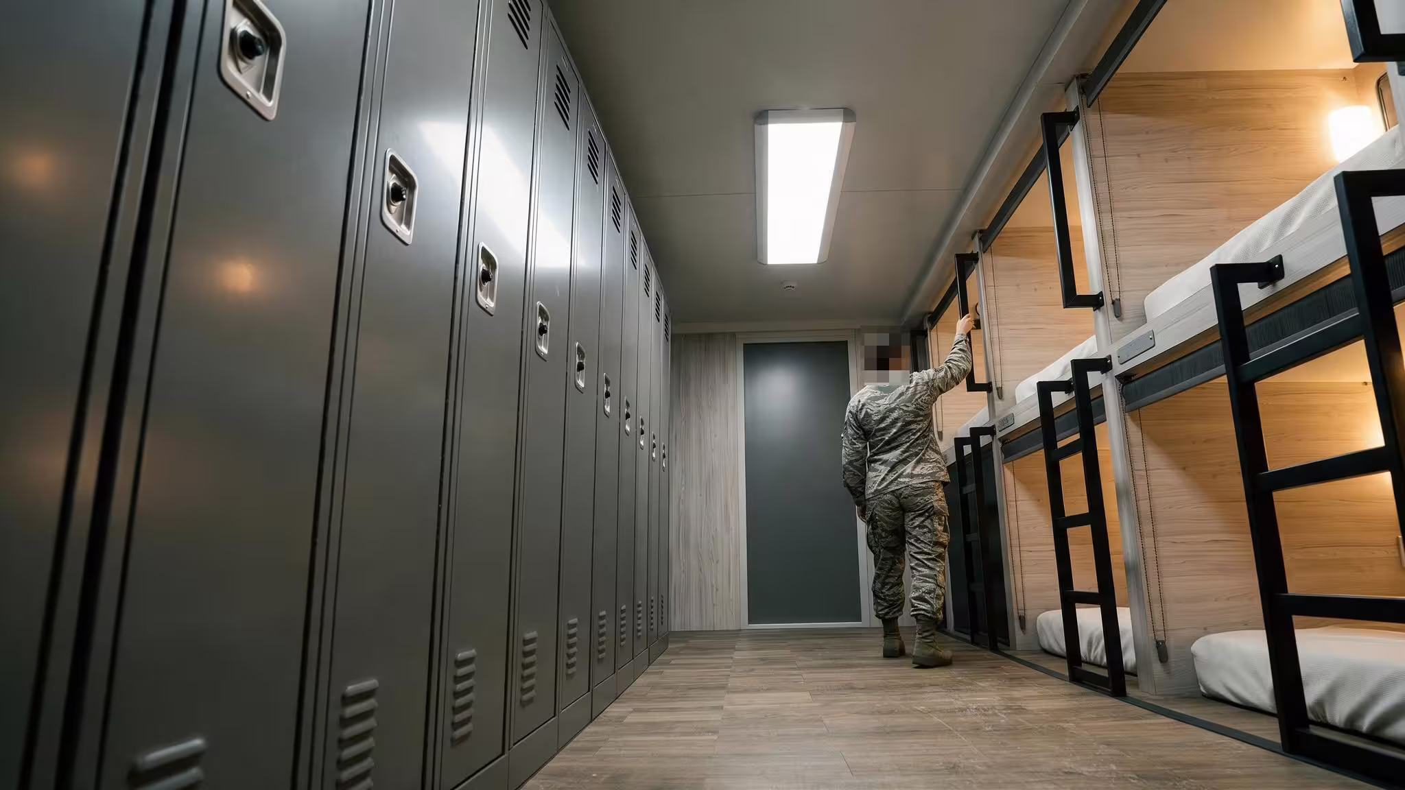 Inside a BunkBox, looking down the central corridor: a full run of tall grey personal lockers on the left, two tiers of berths in pale timber with black ladders and made-up bunks on the right, a bright ceiling panel overhead and a soldier in camouflage reaching up to a berth