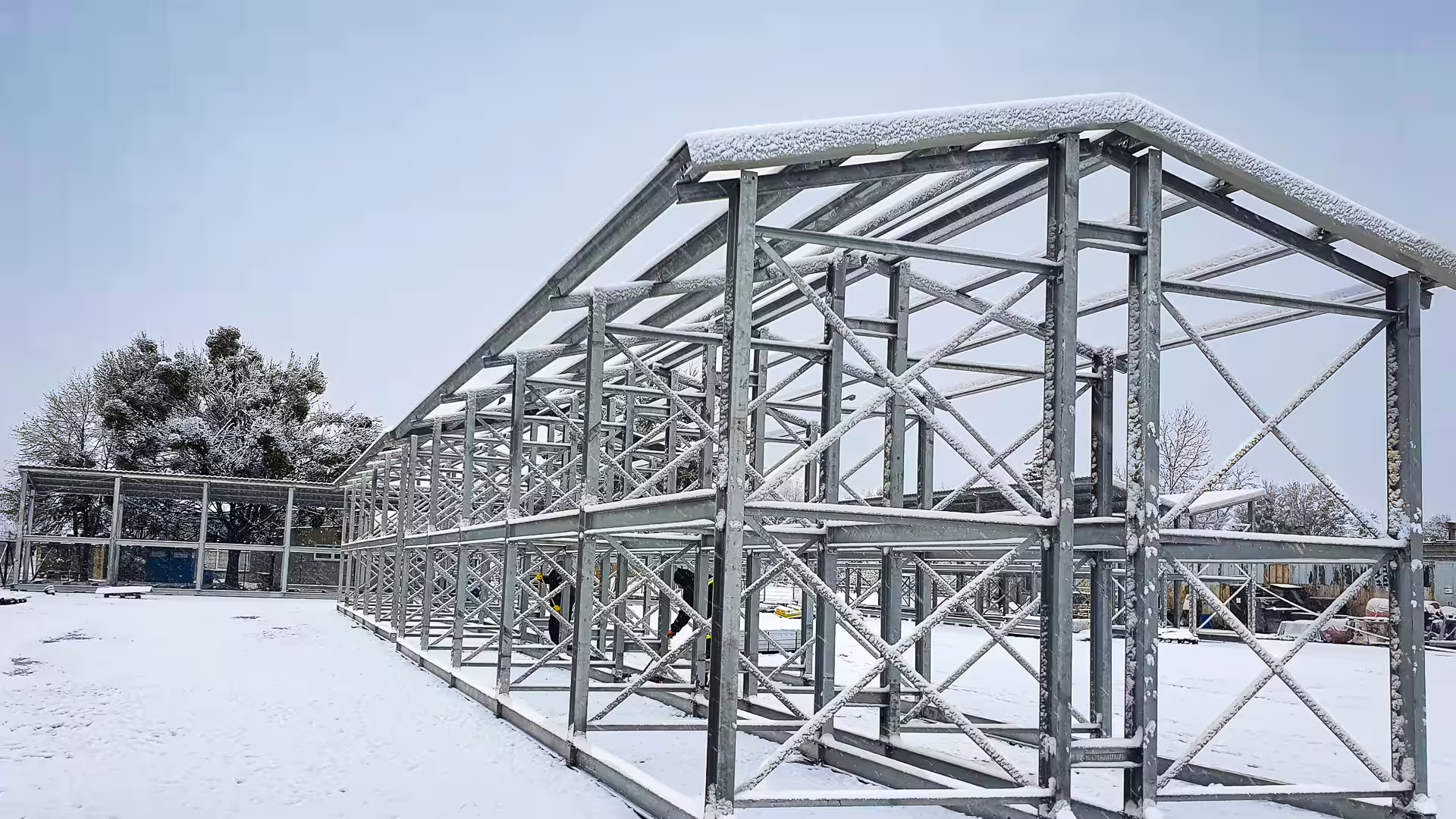 A galvanised CargoRack portal frame standing in a snow-covered yard with its lattice roof trusses and purlins erected and a layer of snow lying across them, a longer roofed run continuing behind it