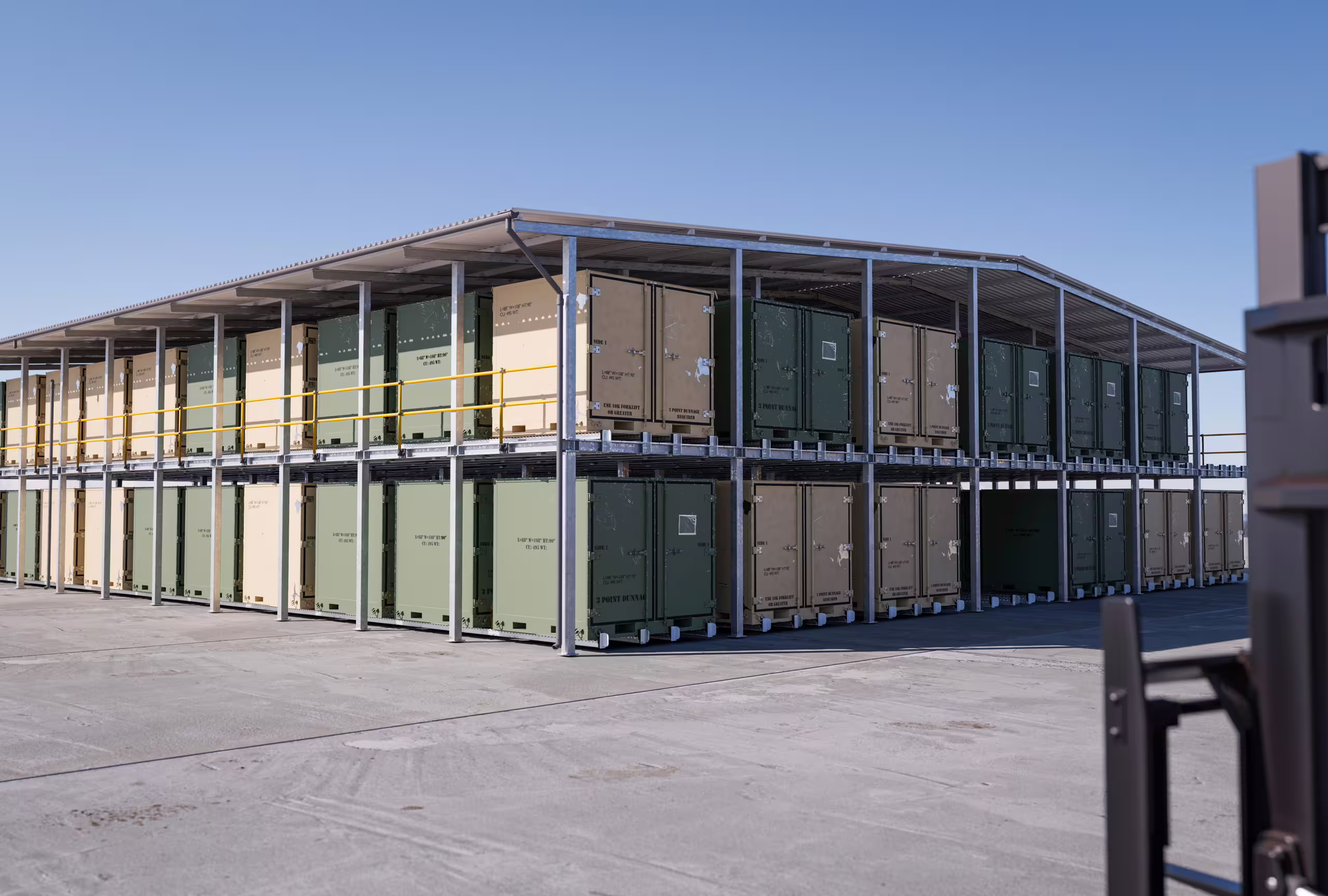 A long two-level CargoRack run on desert hardstanding under a roof, tan and olive containers filling both levels along its length with an access stair at the near end