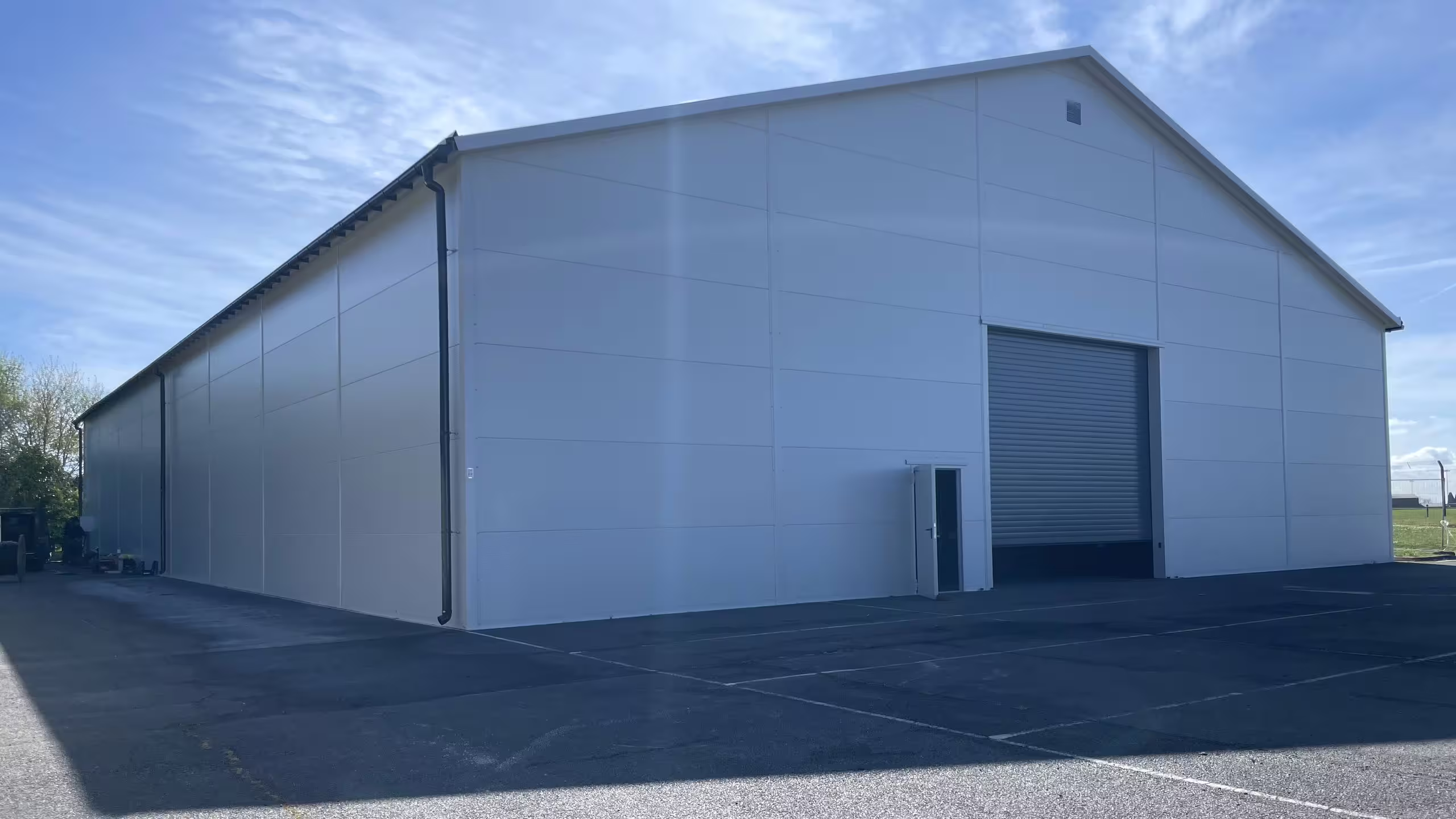 A completed DMTSS shelter on a marked asphalt apron under a clear sky, seen from the corner: white insulated wall panels, a gutter and downpipe along the eaves, a roller-shutter door and a personnel door in the gable end