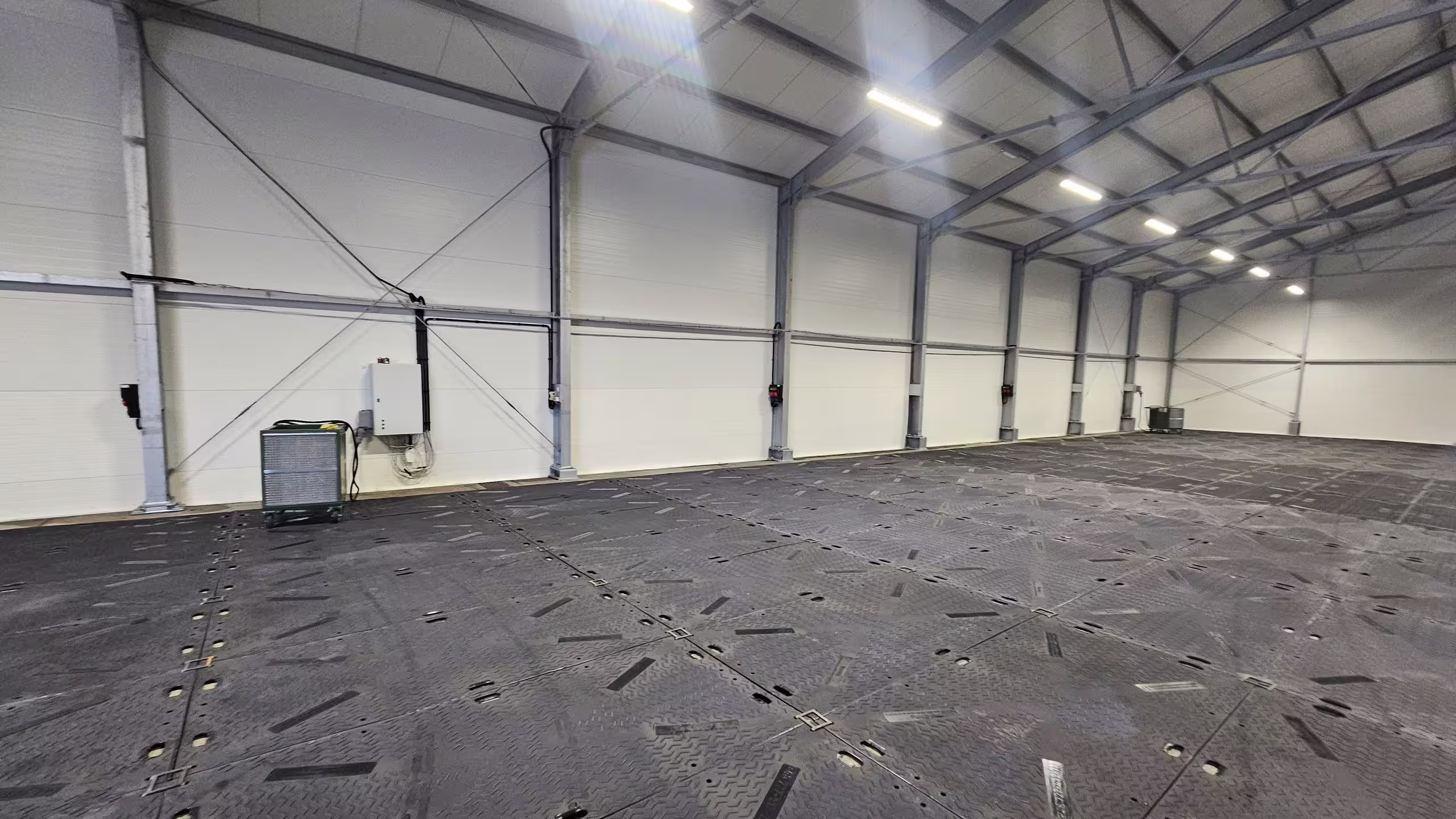 The interior of a DMTSS shelter looking down its length: an unobstructed clear span under a lined roof with continuous lighting runs, the whole floor covered with interlocking composite matting laid over unprepared ground, and a mobile air handler standing against the end wall