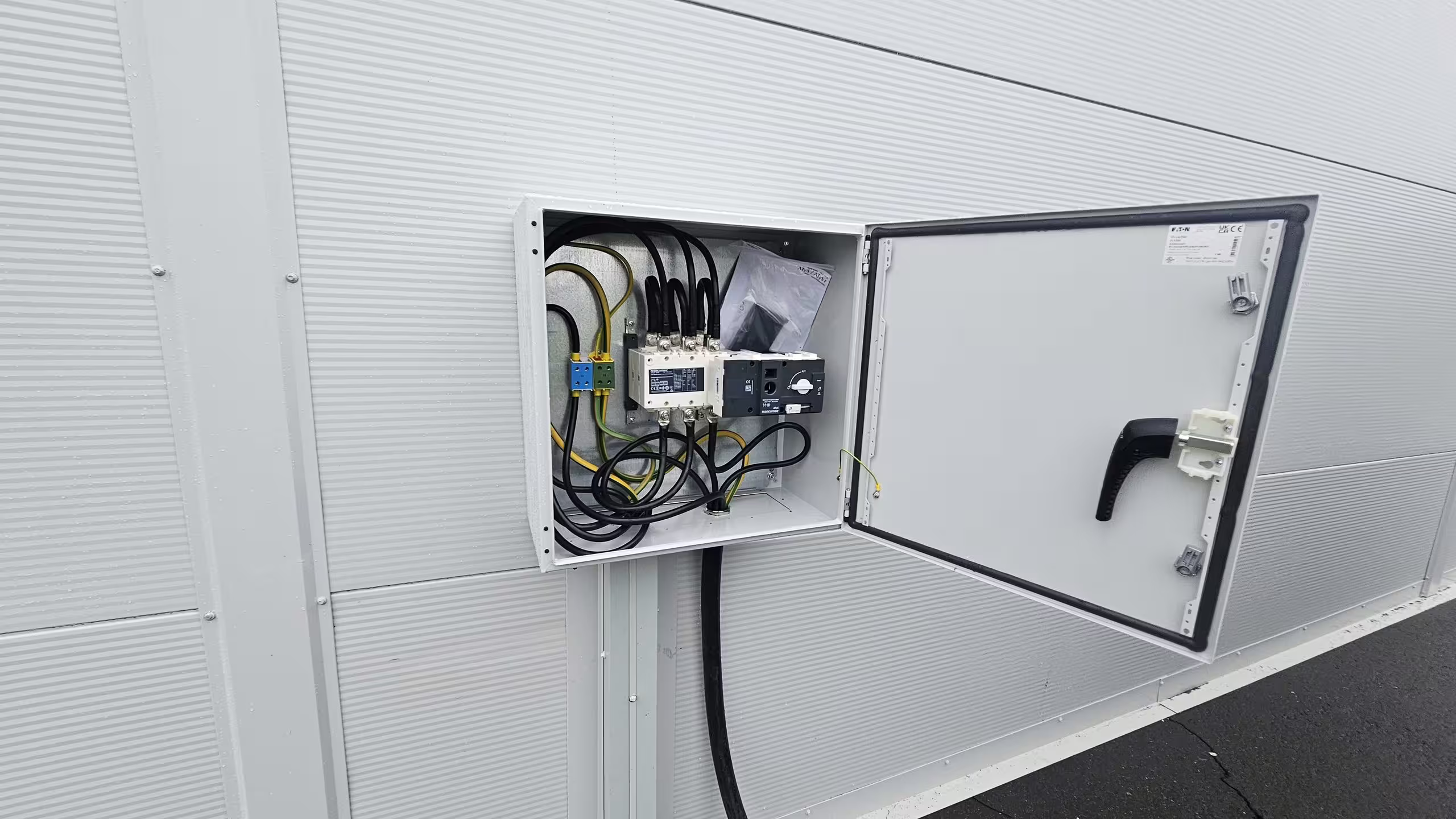 A wall-mounted electrical enclosure on the outside of a DMTSS shelter with its door open, showing a certified mains switch-disconnector and circuit breaker, neatly terminated cabling and an earth bond, fixed to the ribbed insulated wall panel