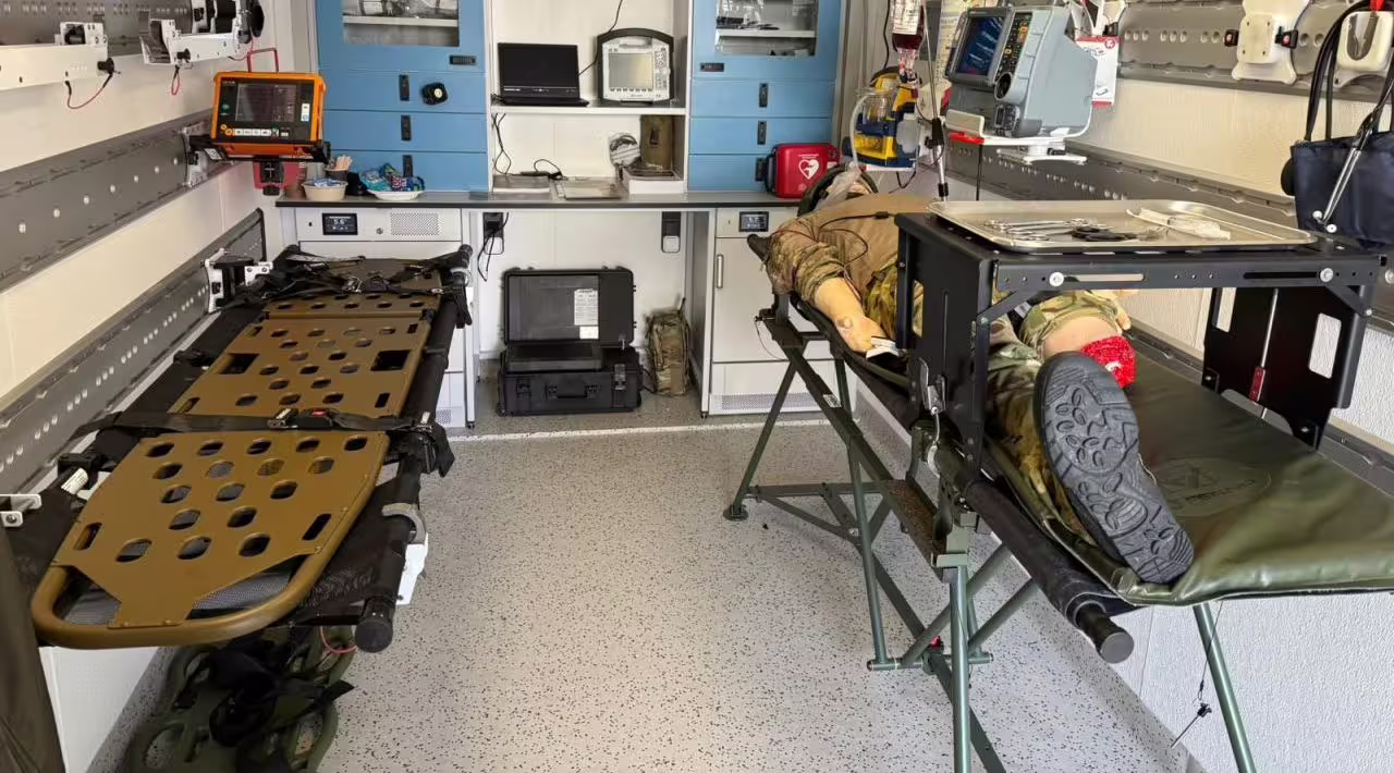 Treatment bay of a fitted FLEXIPOD Clinic with a trauma manikin in combat uniform on a stretcher trolley, an instrument tray over it, a folding litter opposite and a defibrillator on the wall rail