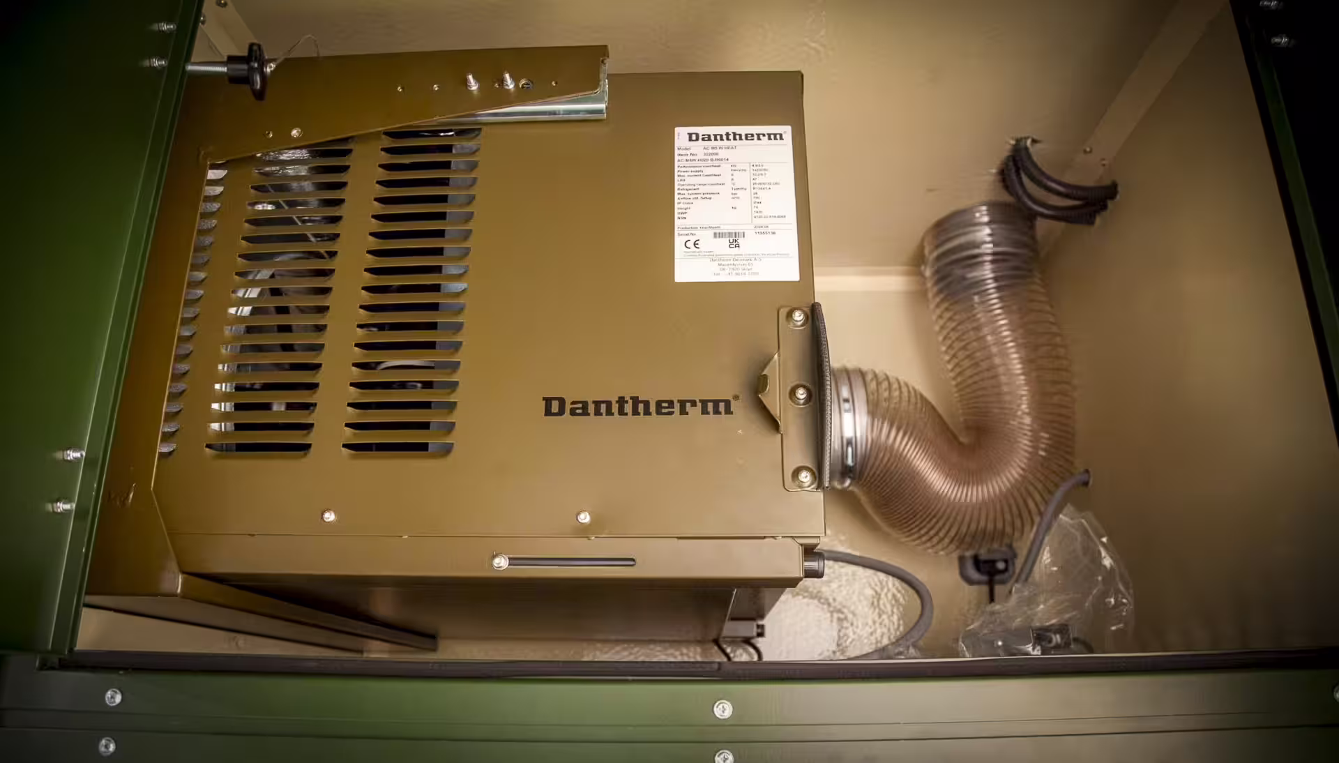 A Dantherm air-handling unit mounted in the clinic's plant bay, its flexible duct connected and the olive access panel folded open beside it