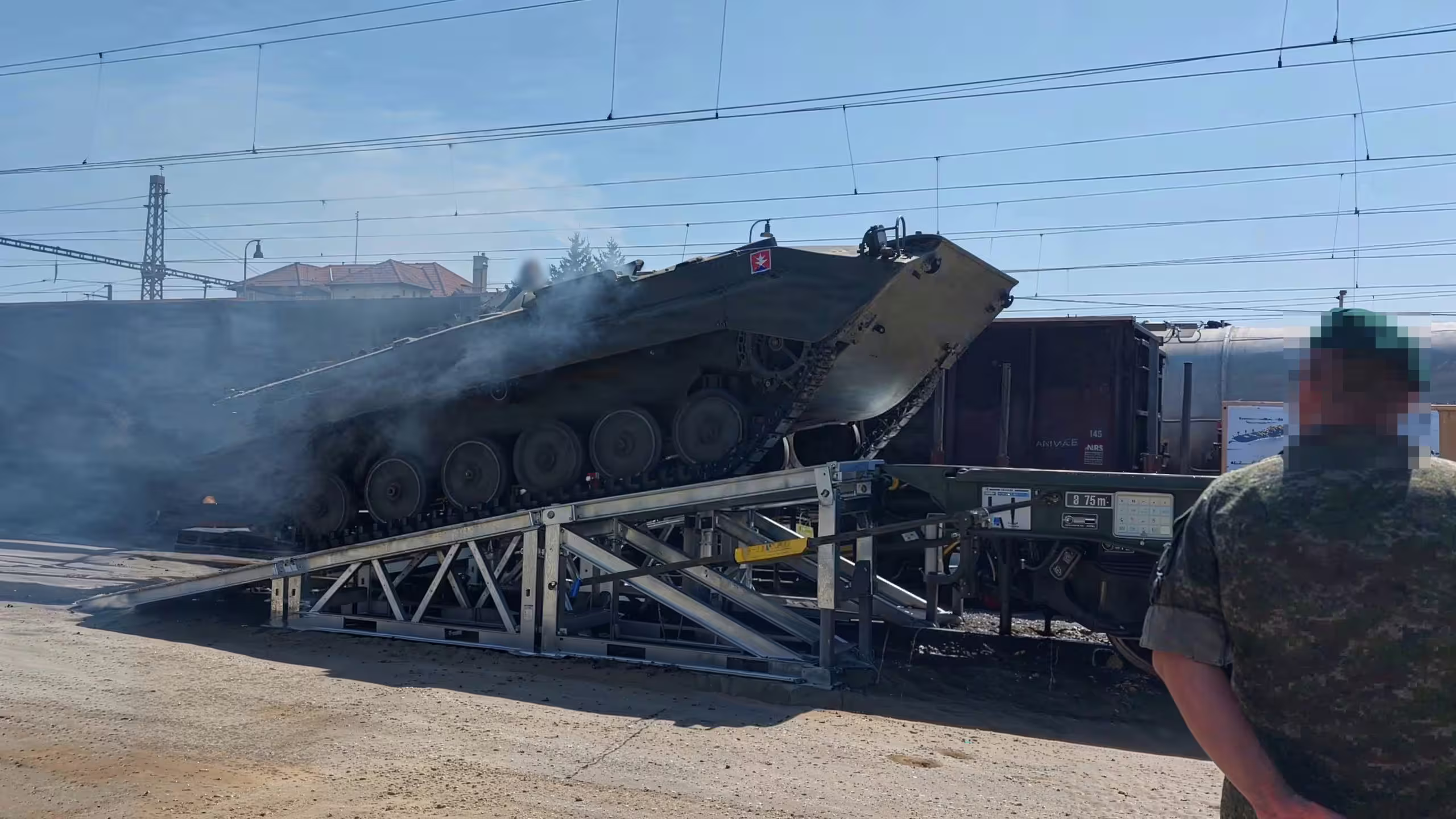 A tracked armoured vehicle driving up a galvanised MUDR-PD90 ramp onto a rail wagon at a railhead, exhaust smoke drifting across the trestle and the deck height set against the wagon floor