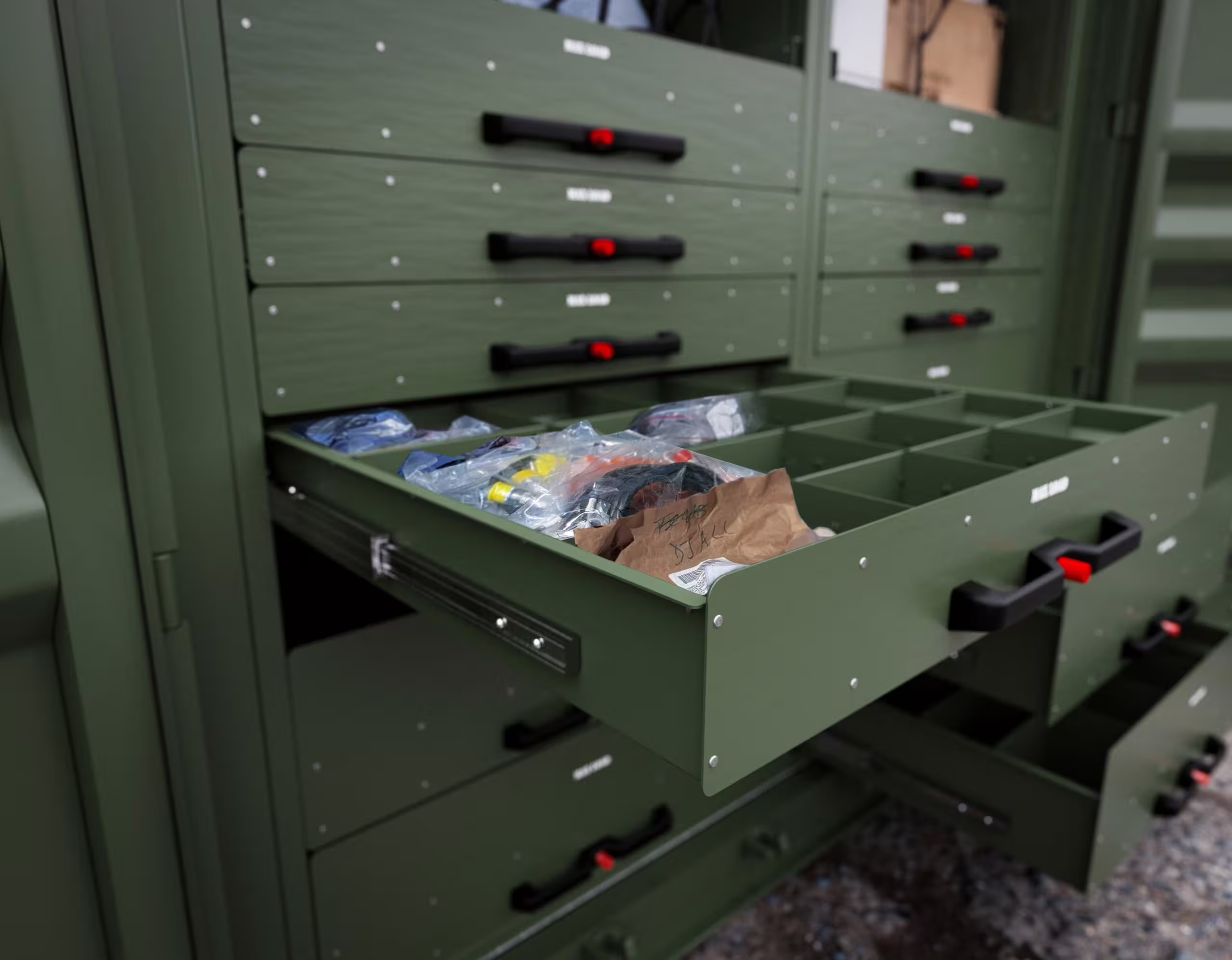 A green Rackbox drawer pulled out on full-extension slides, divided into compartments holding bagged parts and fittings, in a stack of latched drawers inside the container