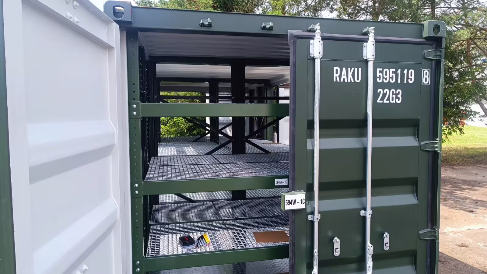 A green 20 ft ISO container with its doors open, three galvanised mesh shelf levels fitted inside and the container's serial and 22G3 type code stencilled on the door
