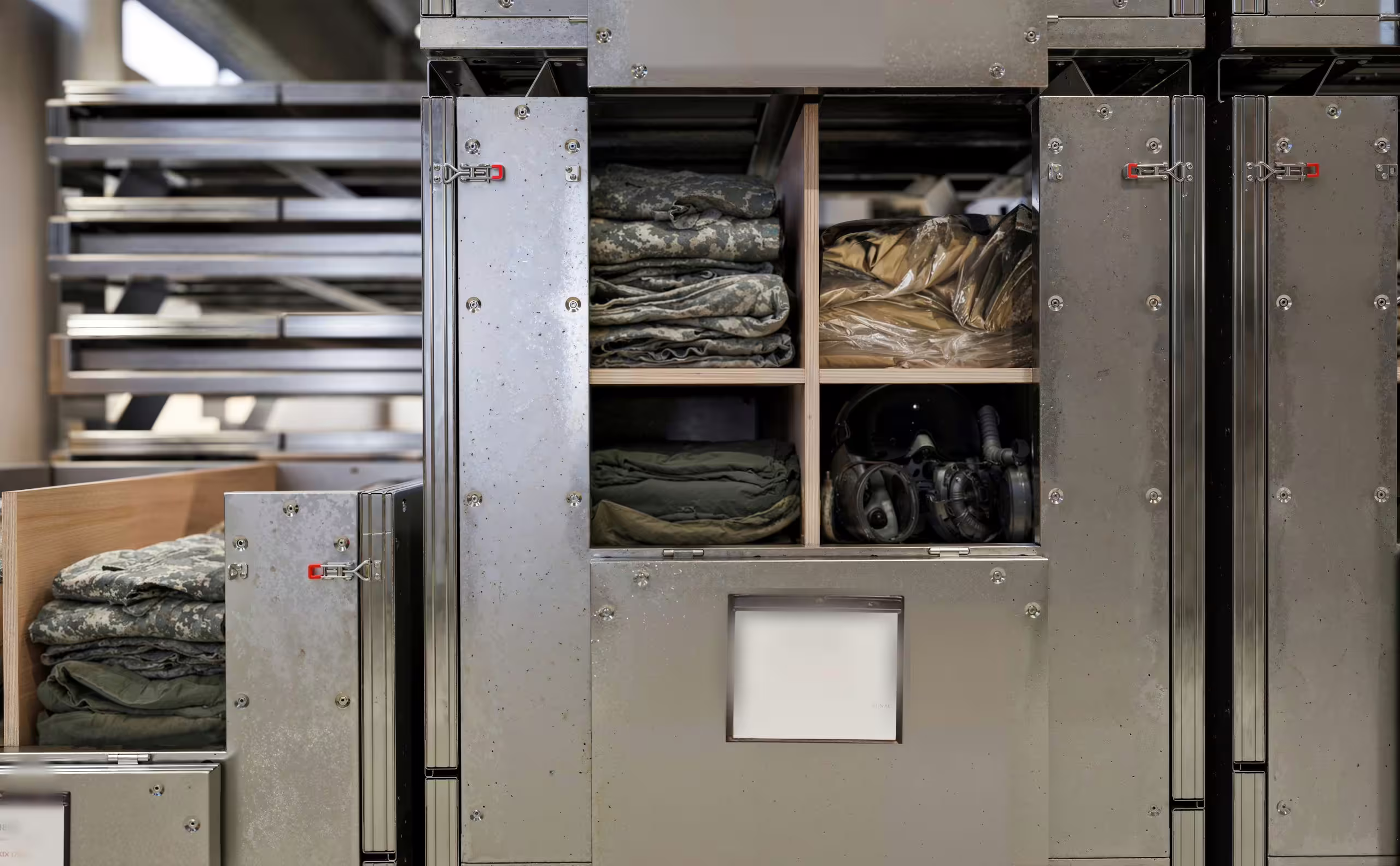 An open SpaceCuber folding bin in a storage bay, its galvanised body divided into compartments holding folded camouflage clothing, packed items and a respirator, with further bins closed alongside
