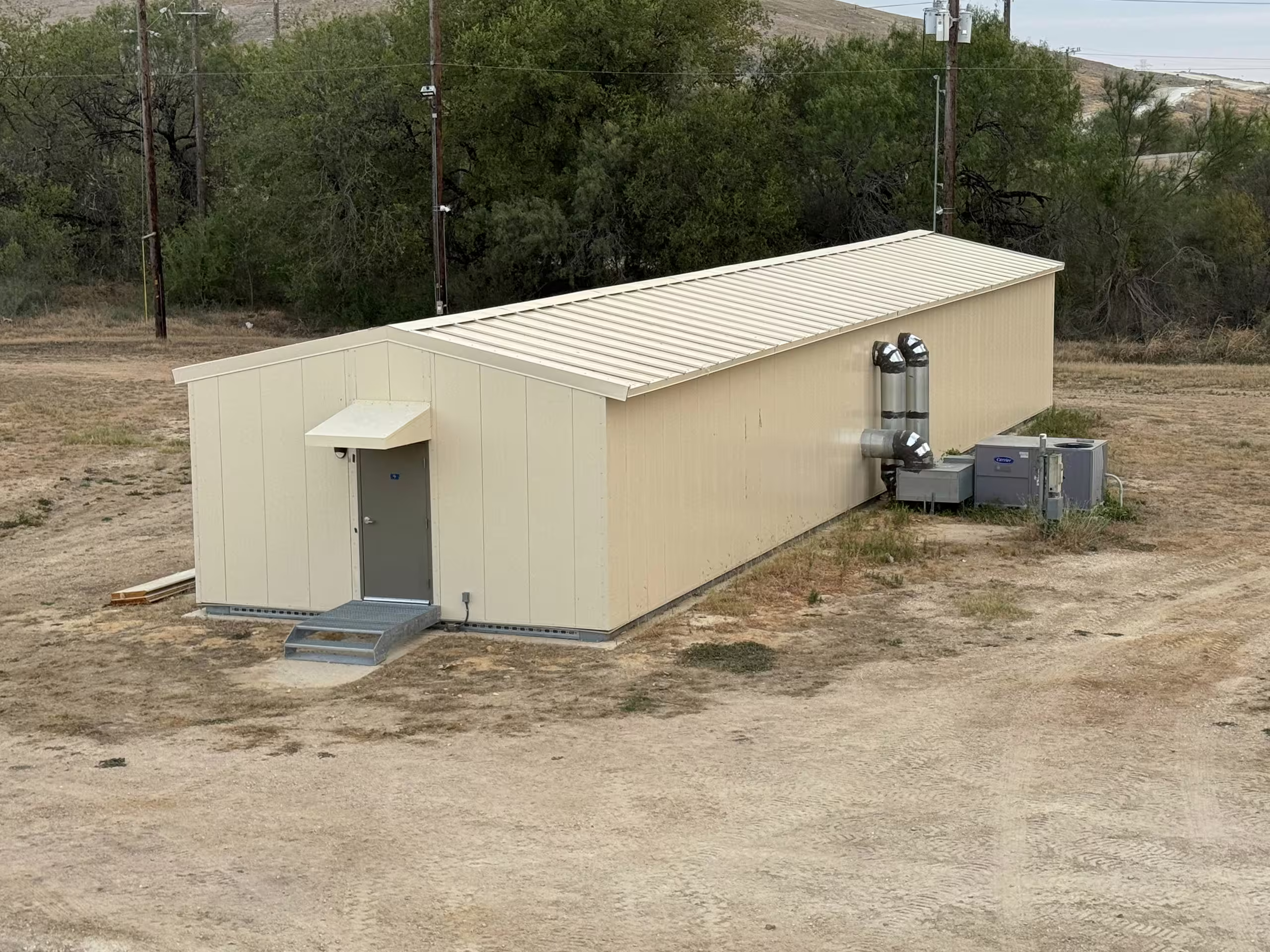 The gable end of a tan Tactical Shelter on dirt hardstanding, with a personnel door, a wall-mounted service unit and conduit runs, scrub and overhead power lines behind