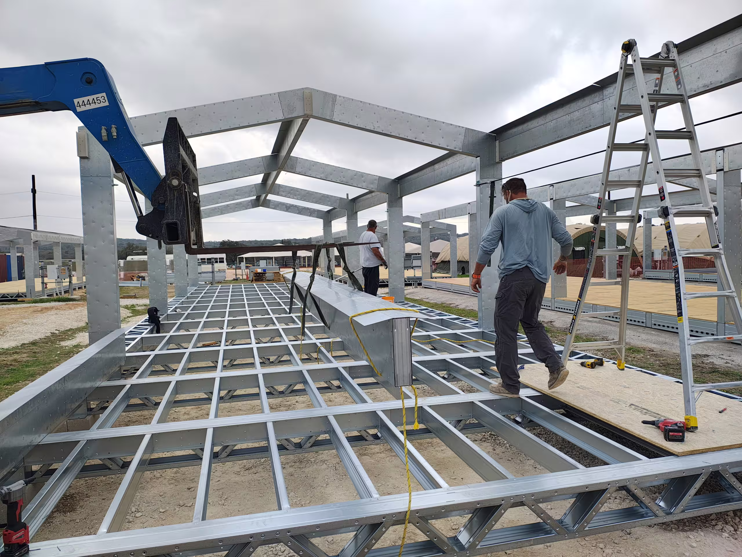 Two fitters working along the open floor structure of a Tactical Shelter on site, lattice floor joists spanning the bay with a step ladder alongside and a telehandler boom overhead