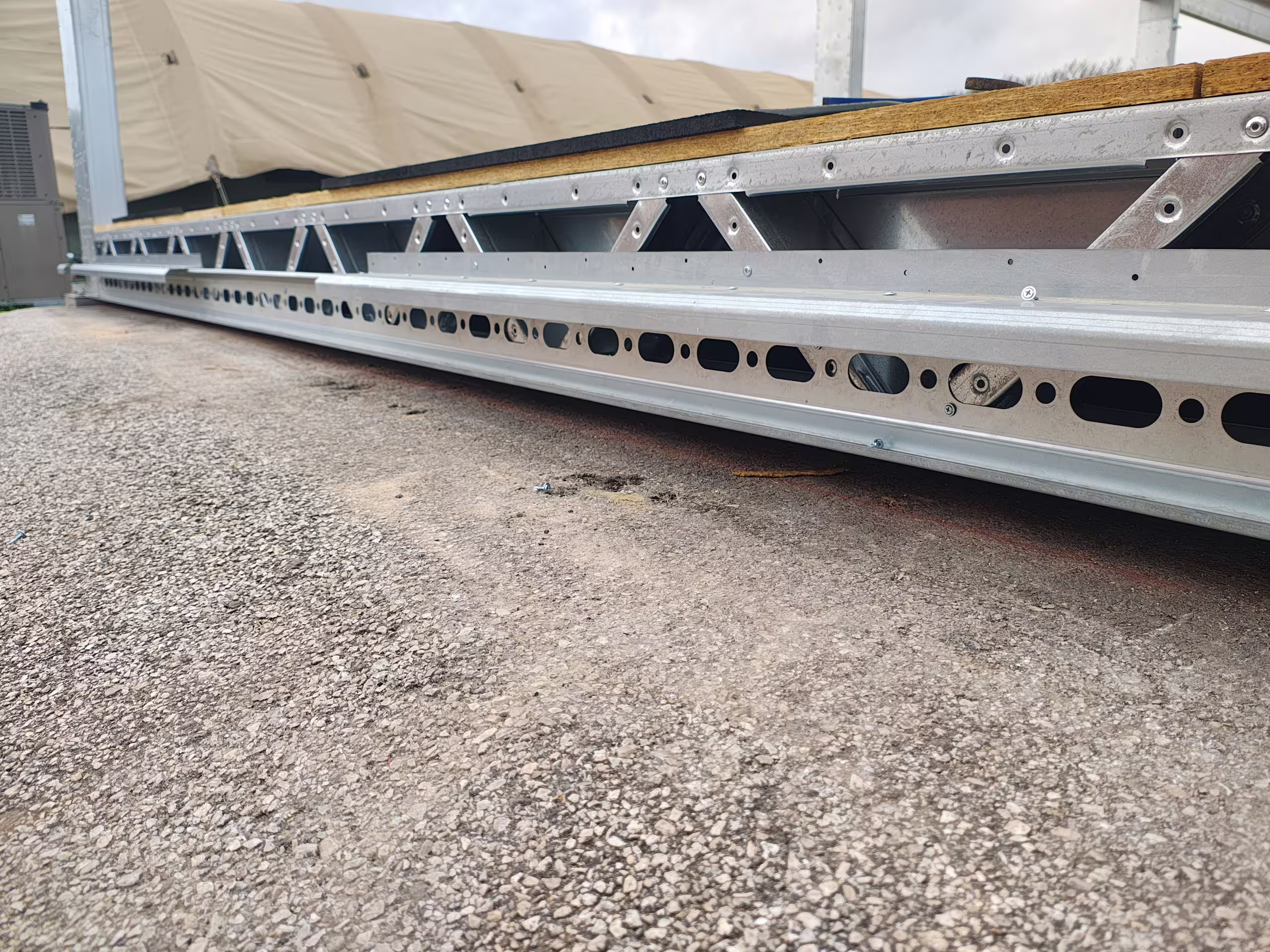 Close-up of Tactical Shelter floor planks and a perforated lattice joist laid on gravel, showing the extrusion profile and bolt holes