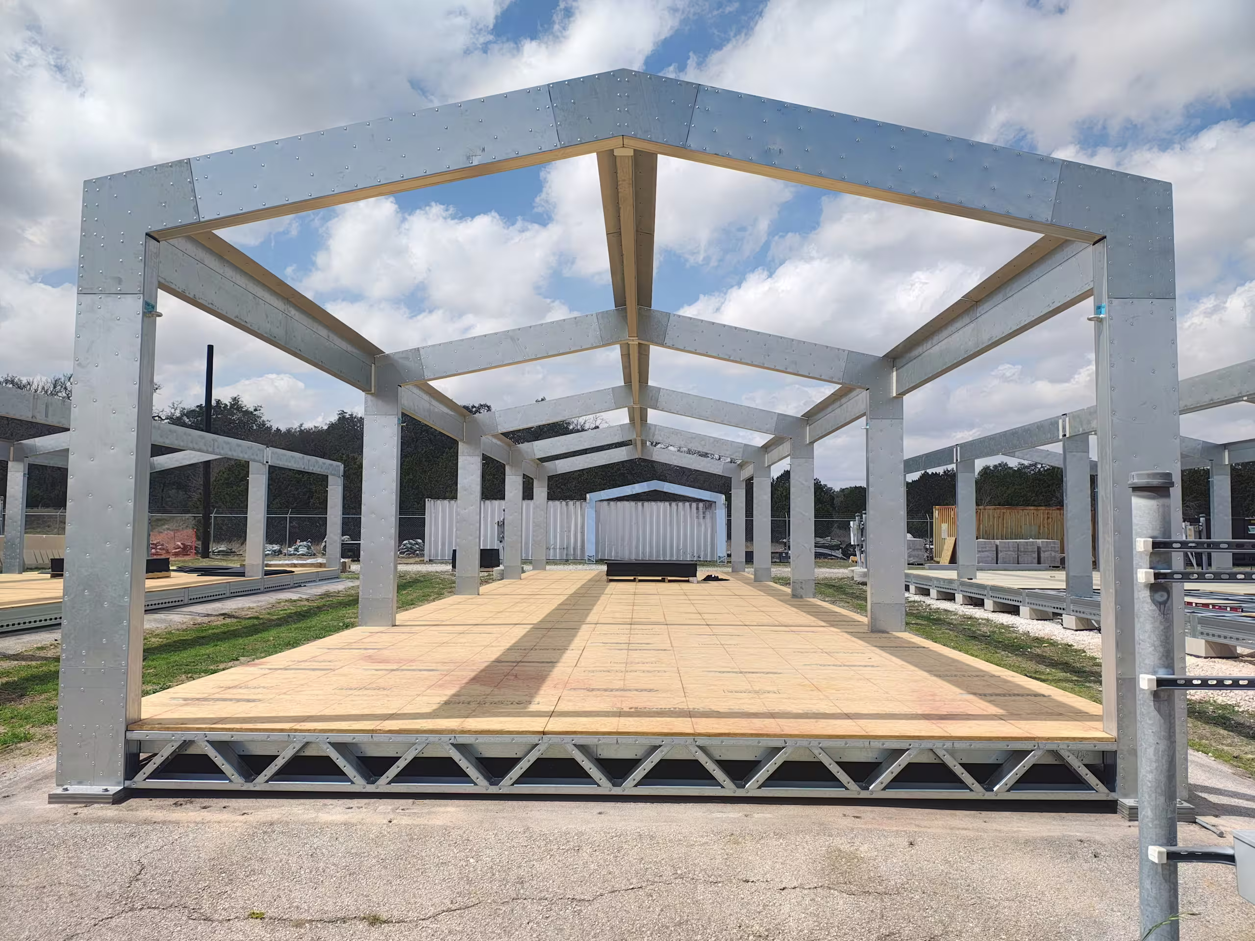 A Tactical Shelter frame erected on its floor deck, portal frames and ridge beams bolted up over board decking, seen from inside the bay under a broken sky