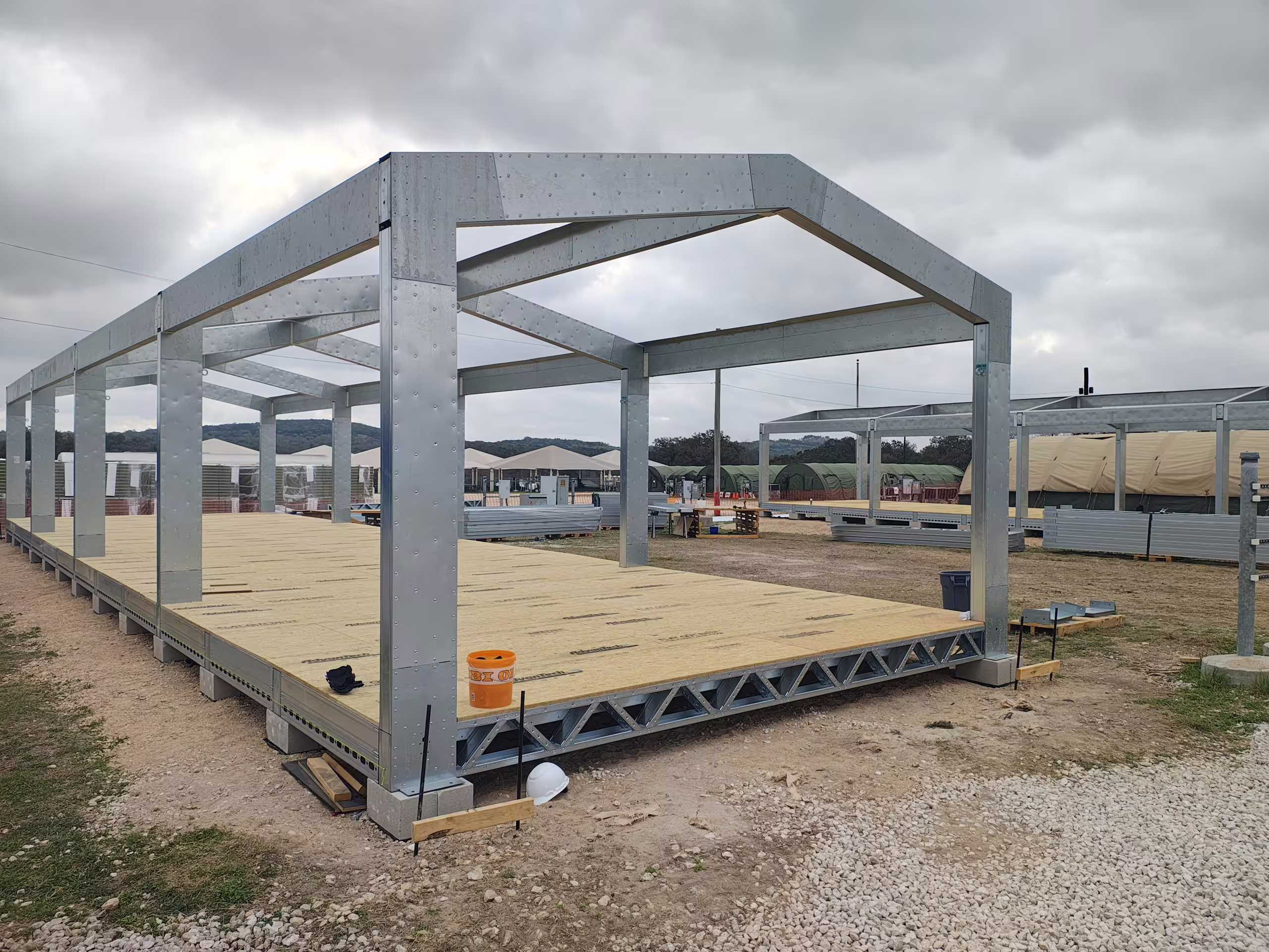 A Tactical Shelter frame standing on gravel with its floor deck laid, bolted portal frames and lattice floor joists, further frames and site buildings behind