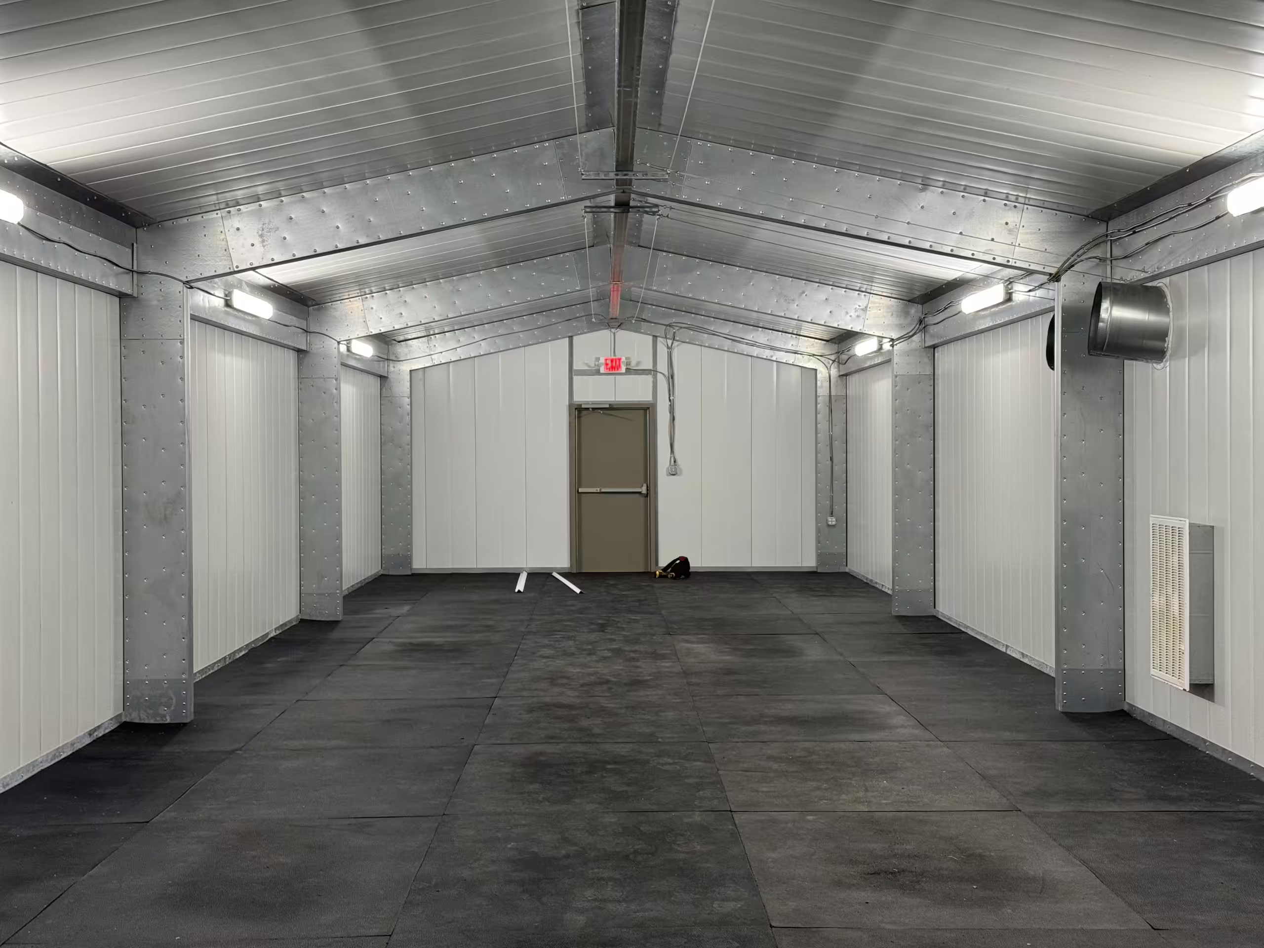 The finished interior of a Tactical Shelter: white insulated wall and ceiling panels, continuous ceiling lighting, a dark floor, a personnel door at the far end and a wall-mounted environmental unit