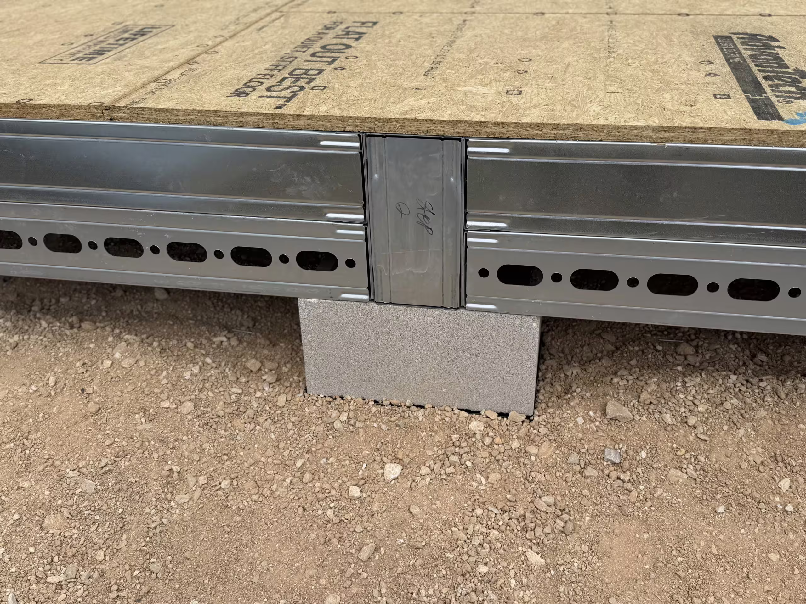 Close-up of the floor edge of a Tactical Shelter under construction: a floor plank sitting on its joist above a packing piece to the gravel below, board decking laid over