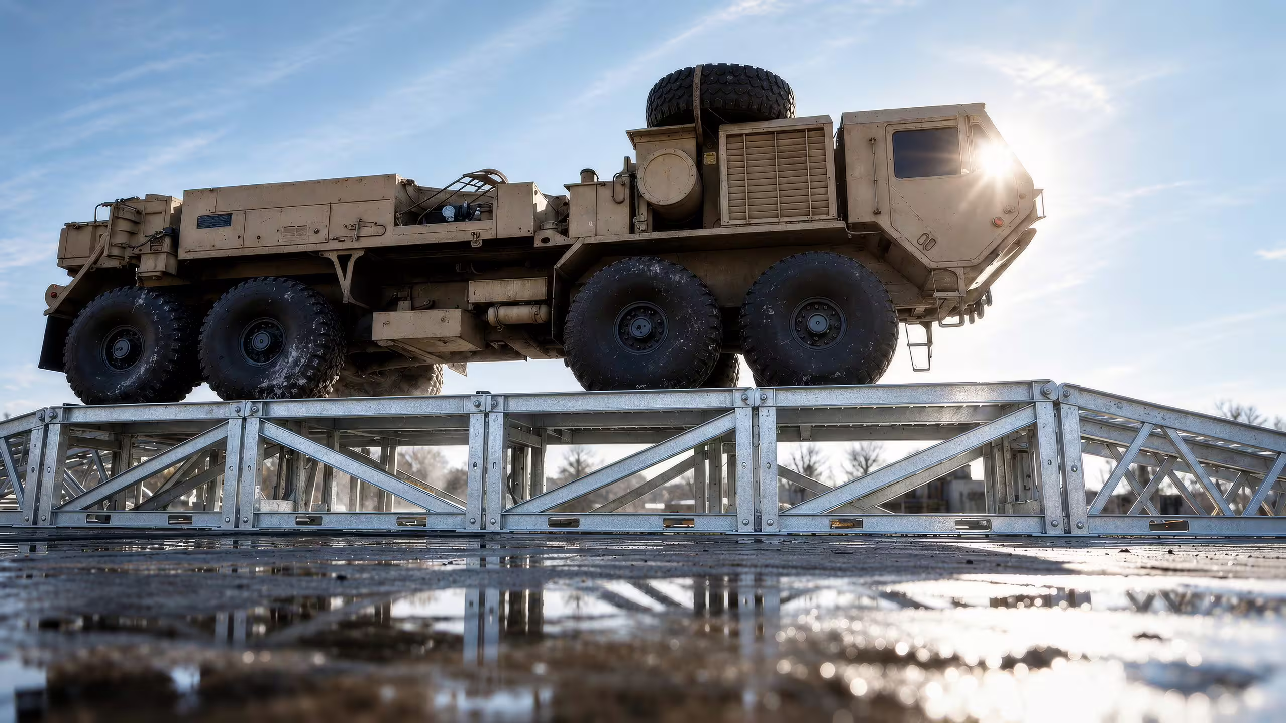 A tan eight-wheeled military truck raised on a galvanised MUDR-PD/WR wash and inspection ramp, the lattice trestle spanning a wet hardstanding with a low sun behind