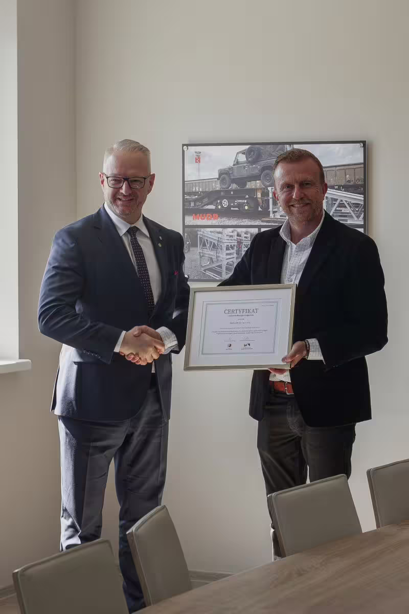 Handover of the WMSSE support decision certificate at the BANAIR plant, two men shaking hands in front of a photograph of a MUDR ramp.