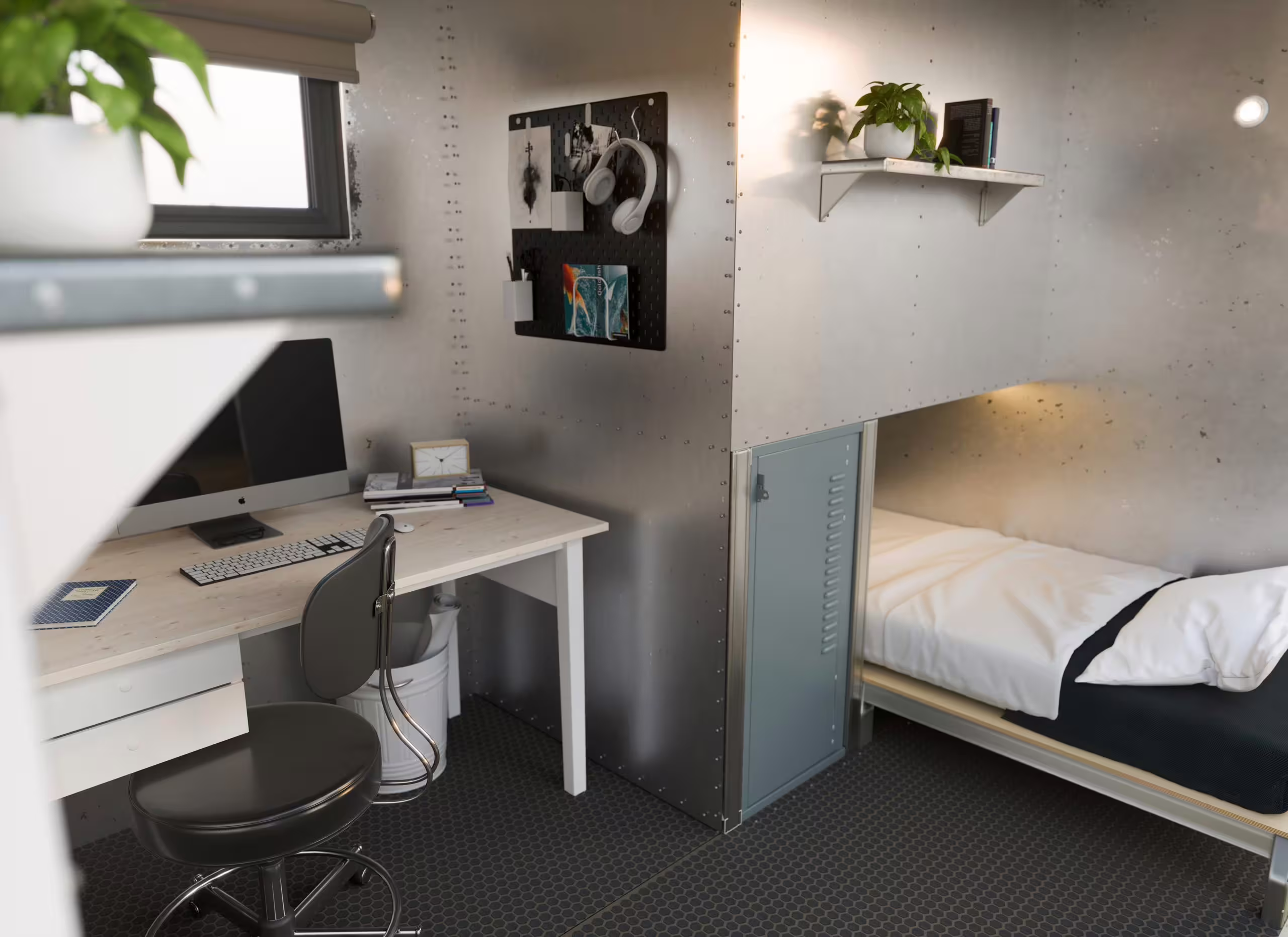 A BunkBox interior configured as a combined office and sleeping space: a desk with a monitor, keyboard and task chair against one wall, a pinboard and shelf of plants above, and a single made bed over a steel locker on the other side