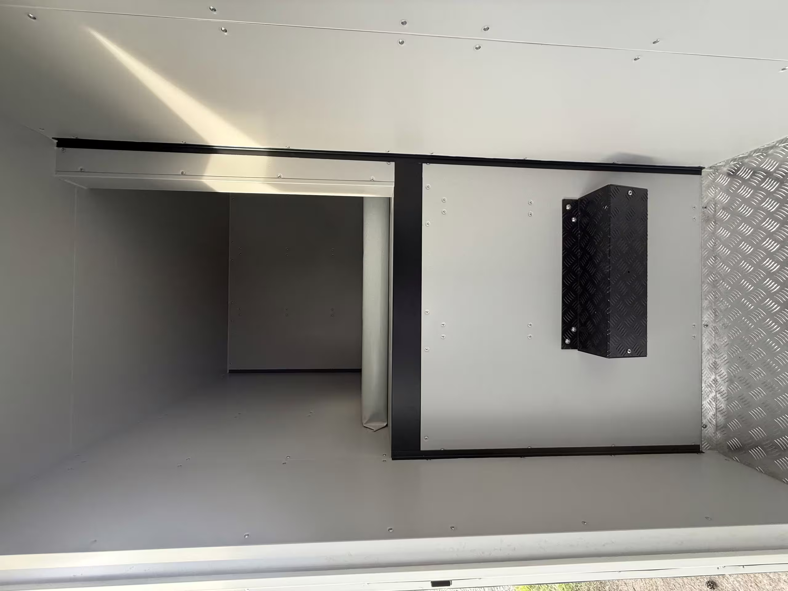 The bare shell of a BunkBox module during build: riveted wall panels, a checkerplate floor and ceiling, an open hatch through the partition and a service box mounted on the wall